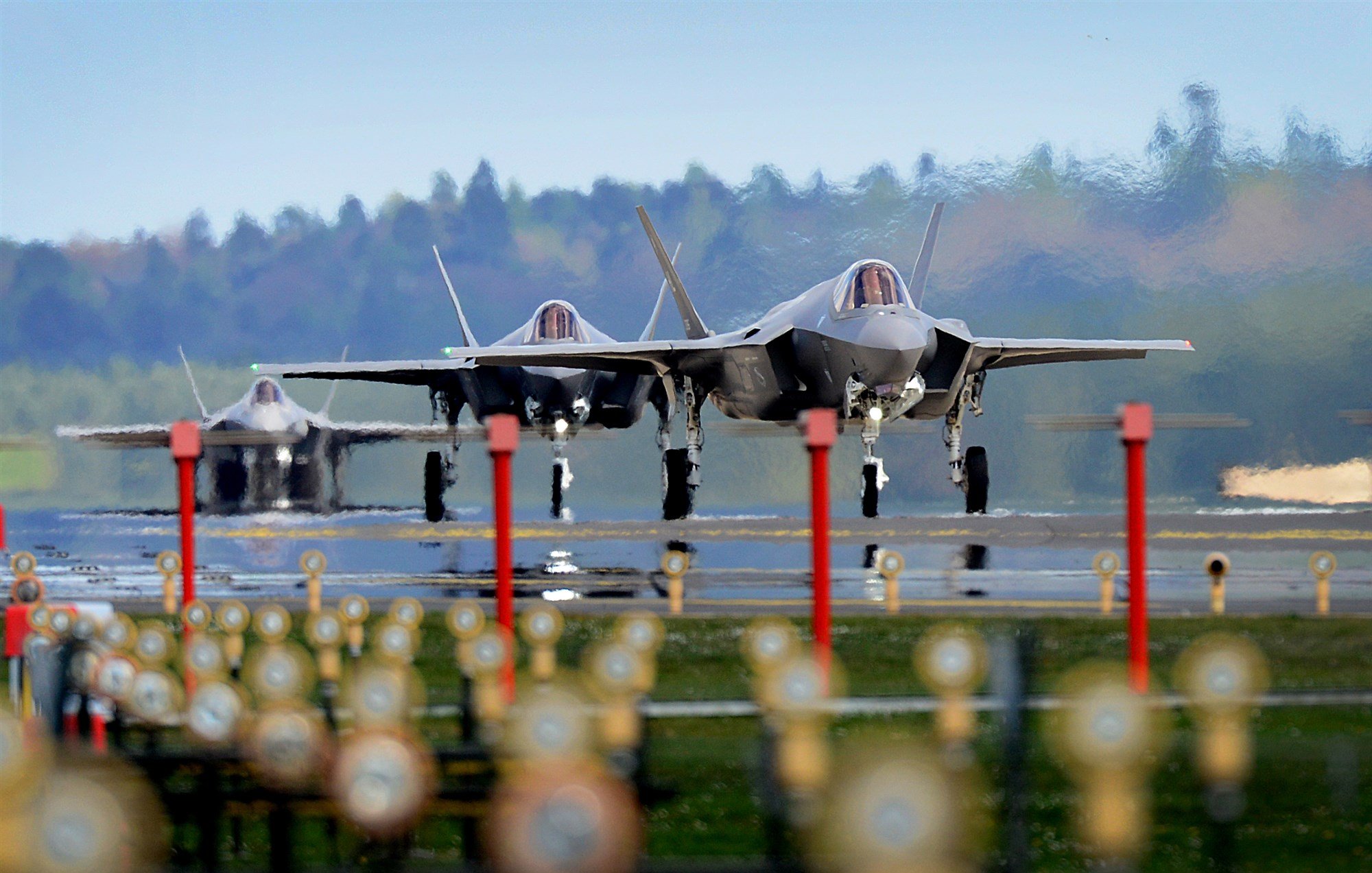 تظهر في الصورة ثلاث طائرات مقاتلة من طراز F-35 تتواجد على مدرج مطار. الطائرات تبدو وكأنها جاهزة للإقلاع، حيث تبرز من الخلف ملامحها الحديثة وتصميمها الأنيق. الأرضية محاطة بمجموعة من الحواجز أو الأعمدة، في حين أن المناظر الطبيعية في الخلفية تظهر أشجارًا ومساحات خضراء تحت سماء زرقاء. تُعطي الصورة انطباعًا عن القوة والتقنية المتقدمة في مجال الطيران العسكري.