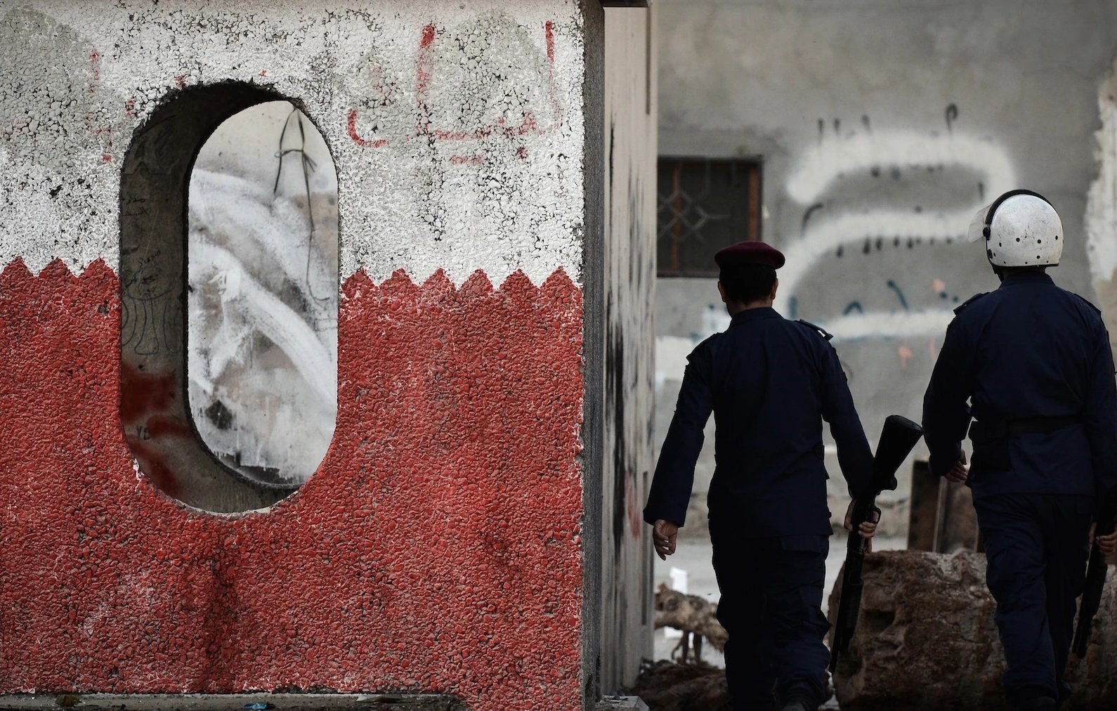 The image shows two police officers walking away from a wall that has a distinctive design. The wall features a rough texture with a circular opening and a color scheme that includes red and white. The background suggests an urban environment, with graffiti and signs of wear on the walls. The officers are dressed in dark uniforms and appear to be carrying weapons, indicating a presence of law enforcement in a possibly tense or unrested area.