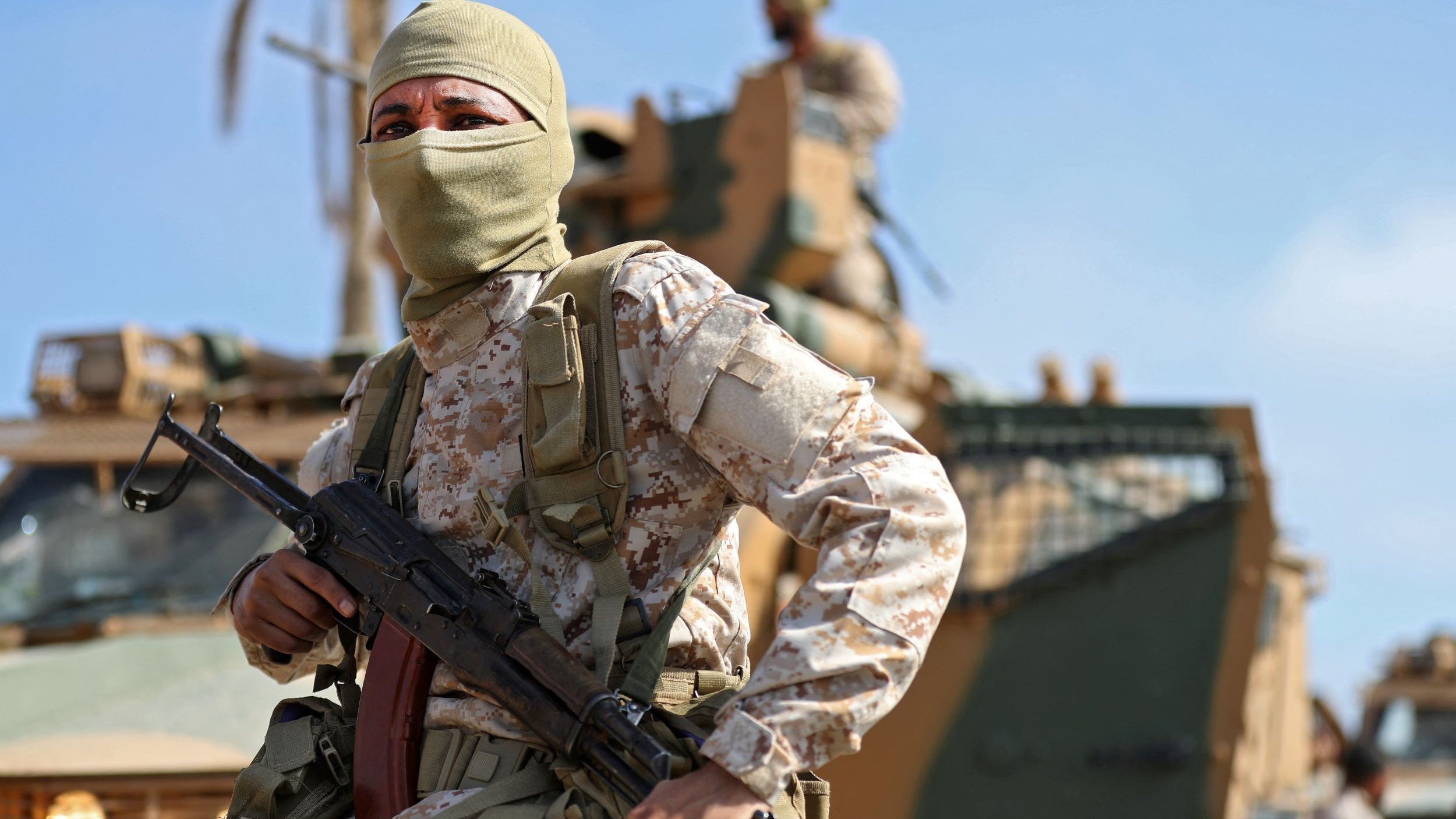 در این تصویر، یک سرباز مسلح با پوشش صورت، در حال ایستادن در پیش‌زمینه قرار دارد. او یونیفورم نظامی و کلاهی به سر دارد و یک اسلحه در دستش است. در پس‌زمینه، یک خودروی نظامی دیده می‌شود و تعدادی از سربازان دیگر در حال فعالیت هستند. آسمان آبی و آب و هوای روشن، صحنه را روشن می‌کند. این تصویر حس قدرت و دقت نظامی را به تصویر می‌کشد.