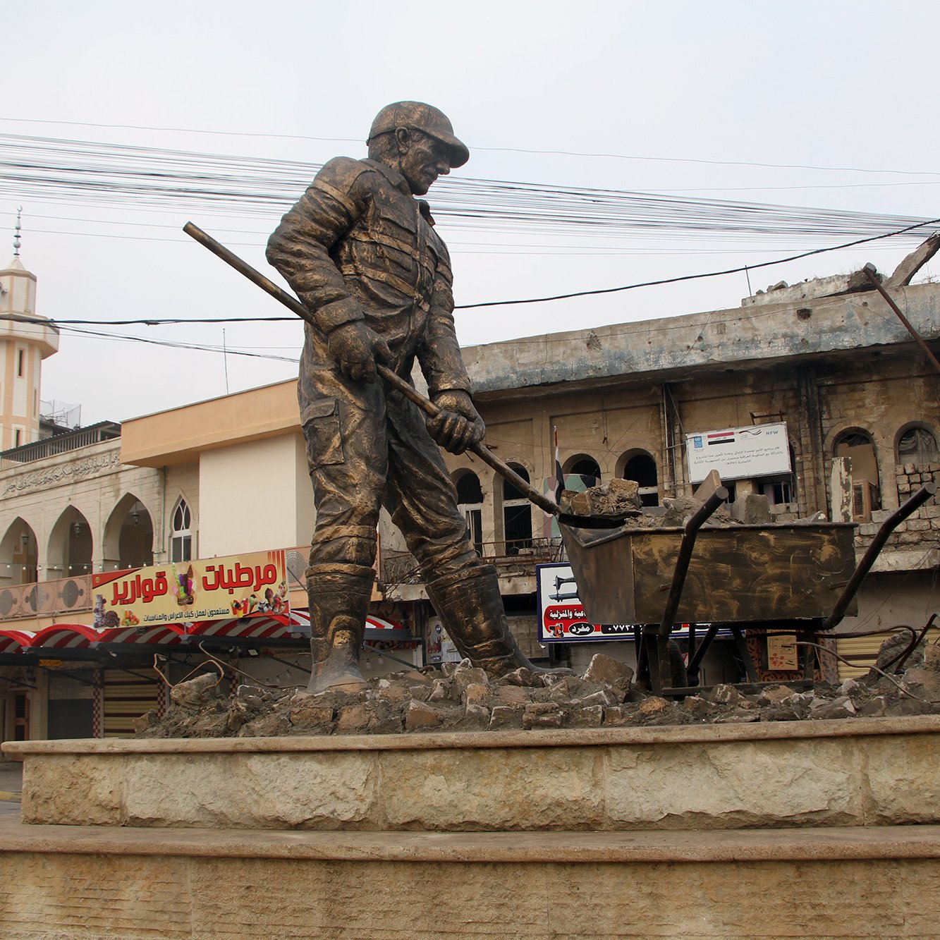 L'immagine mostra una statua di un operaio che lavora, con una pala in mano e una carriola accanto a lui. Il monumento è situato in una zona urbana, caratterizzata da edifici danneggiati e in rovina sullo sfondo. La scena trasmette un senso di resilienza e dignità, riflettendo la vita e il lavoro in un contesto difficile. L'atmosfera è un po' desolata a causa delle strutture danneggiate, mentre la statua rappresenta il duro lavoro e l'impegno quotidiano delle persone.