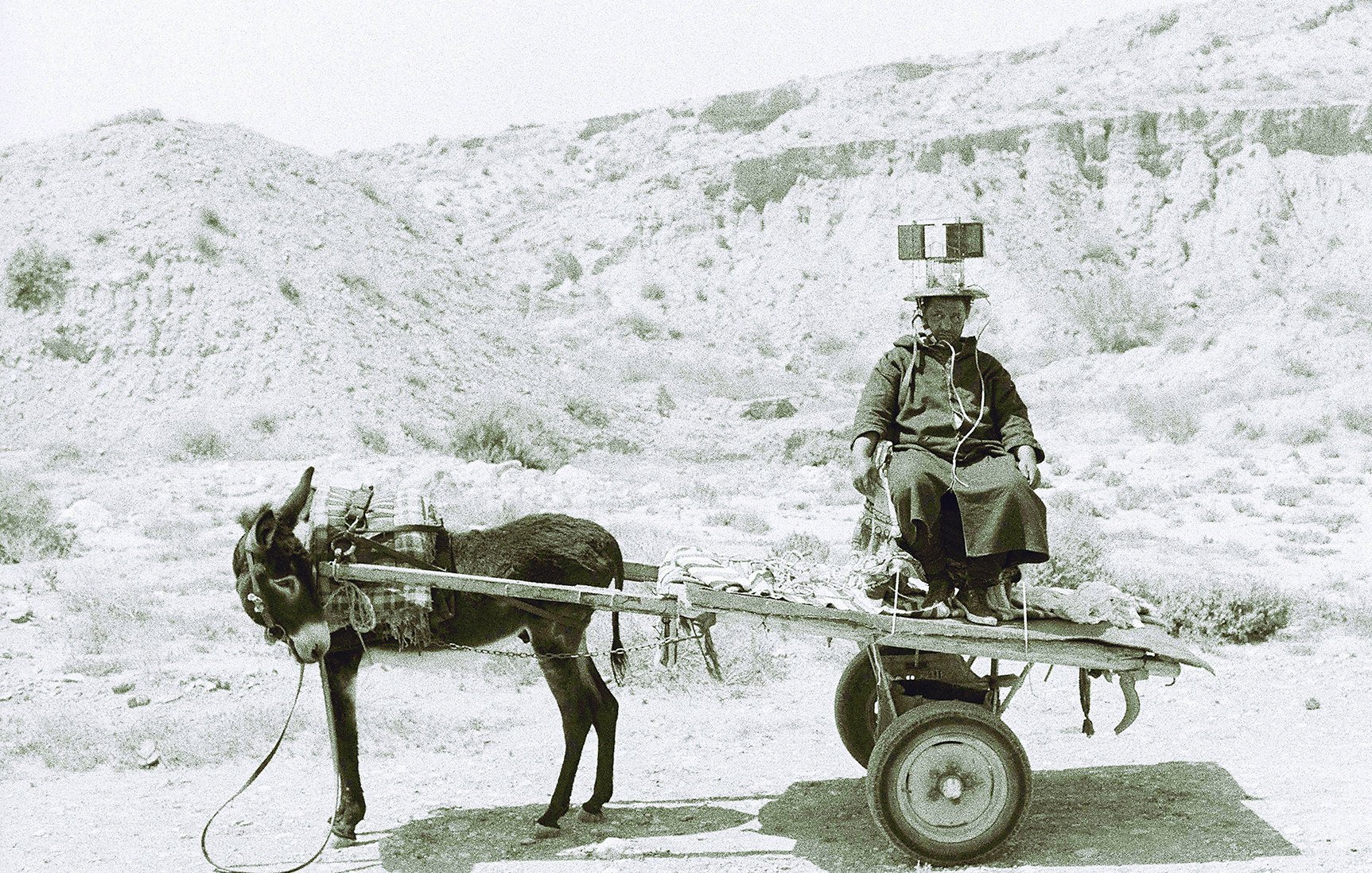 L'image montre une scène en noir et blanc d'un homme assis sur un chariot tiré par un âne. L'homme porte des vêtements traditionnels et semble se diriger quelque part, tandis que l'âne est attaché à l'avant du chariot. L'arrière-plan présente un paysage aride et désertique, avec des collines ou des montagnes à l'horizon. L'atmosphère de la photographie évoque un sentiment de simplicité et de vie rurale.