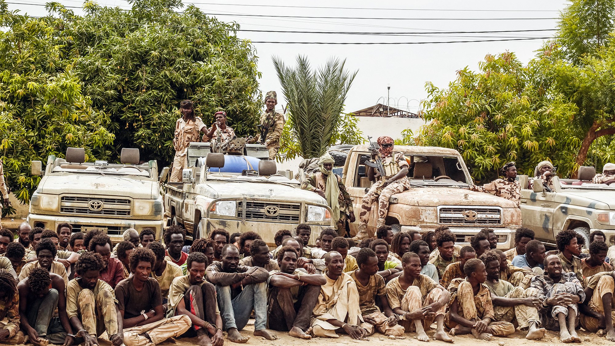 L'image montre un groupe de personnes assises au sol, semblant fatiguées et dans des vêtements usés, tandis qu'elles sont entourées de plusieurs véhicules tout-terrain. À l'arrière-plan, on peut voir des arbres et des plantes, suggérant un environnement rural. Certains individus semblent garder les véhicules, et l'atmosphère générale donne une impression de tension ou de détresse.