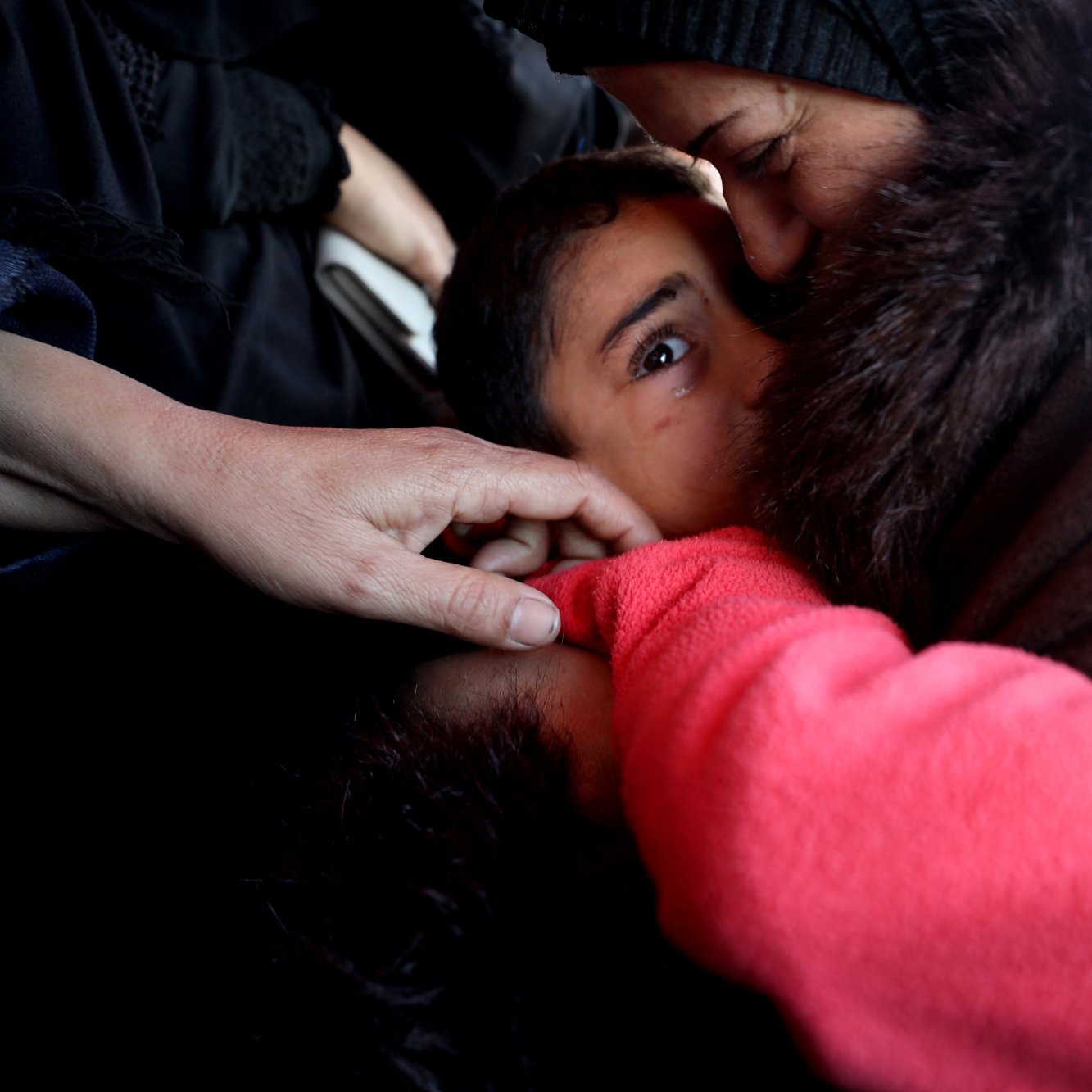 The image depicts a close interaction between an adult woman and a child. The woman is gently kissing the child on the forehead, while the child looks directly at the camera with a serious expression. There are hands visible in the foreground, suggesting that others are present nearby. The overall atmosphere conveys a sense of warmth and tenderness despite the child's intense gaze. The setting appears to be intimate, possibly capturing a moment of care and connection in a challenging situation.