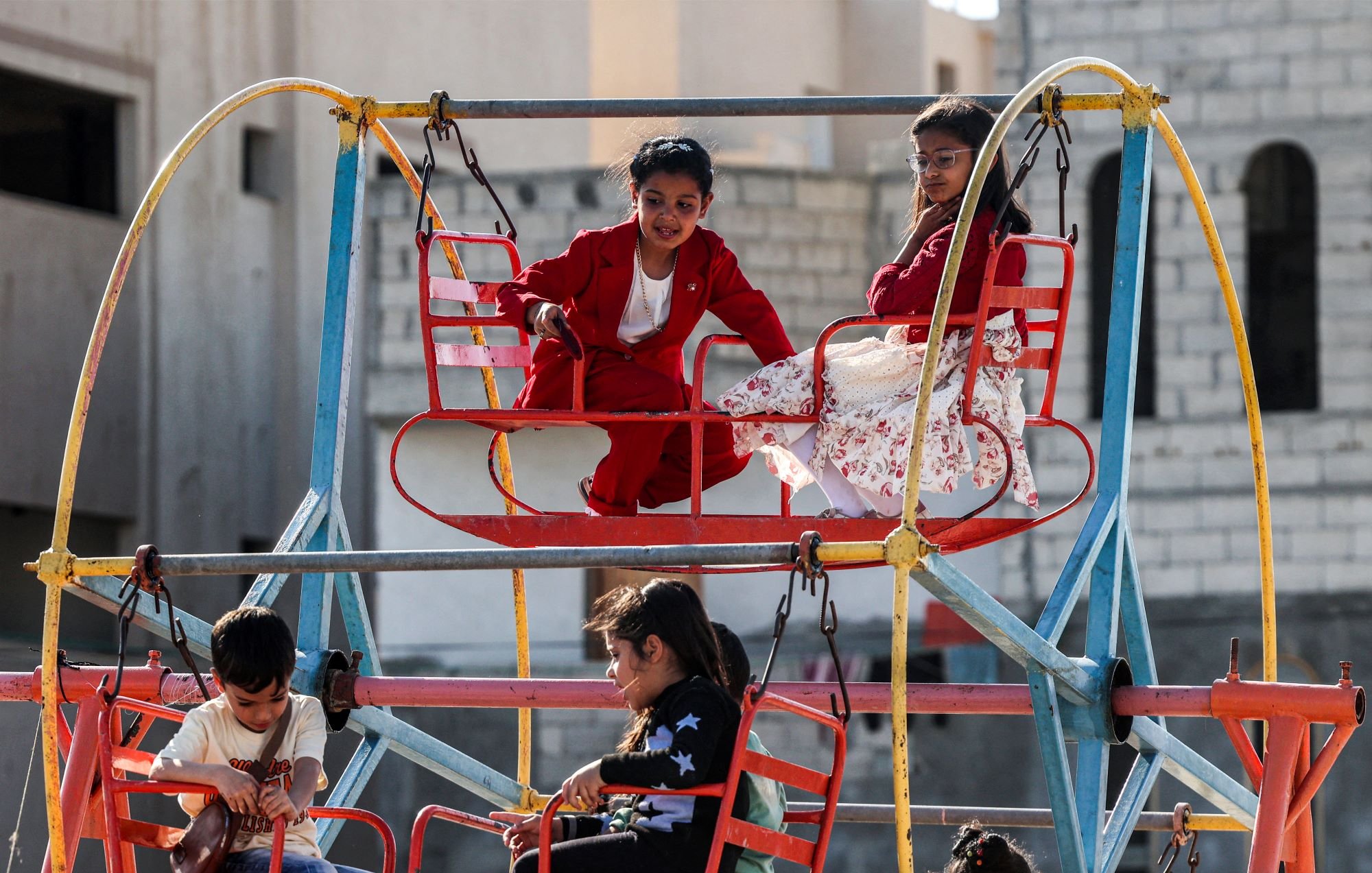 L'image montre des enfants jouant sur une grande structure de jeux colorée. On voit des balançoires fixées à une plateforme, dont certains enfants sont assis. Les enfants affichent des expressions joyeuses, profitant de leur temps de jeu. La scène se déroule dans un espace ouvert, avec des bâtiments en arrière-plan. Les vêtements des enfants sont variés, ajoutant de la vivacité à l'ensemble. L'atmosphère semble joyeuse et familiale.