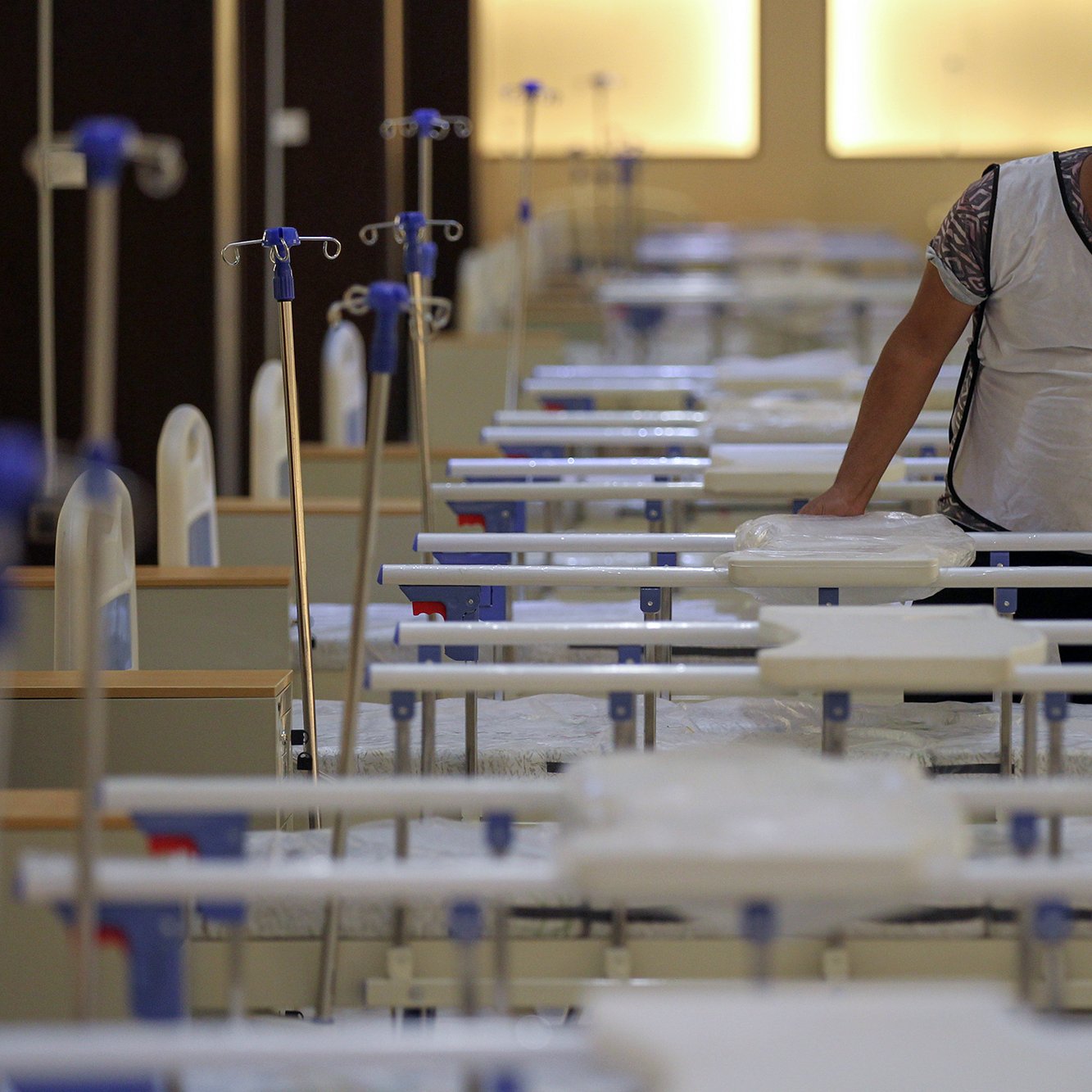 در این تصویر، یک مرد در حال کار در یک فضای بیمارستانی یا درمانگاهی دیده می‌شود. او یک ماسک به صورت دارد و به تخت‌هایی که در ردیف‌های مرتب قرار دارند، توجه می‌کند. تخت‌ها با ملحفه‌های سفید پوشیده شده‌اند و تعدادی از آن‌ها به دستگاه‌های اکسیژن متصل هستند. نور محیط ملایم است و فضا به نظر تمیز و سازمان‌دهی شده می‌رسد.