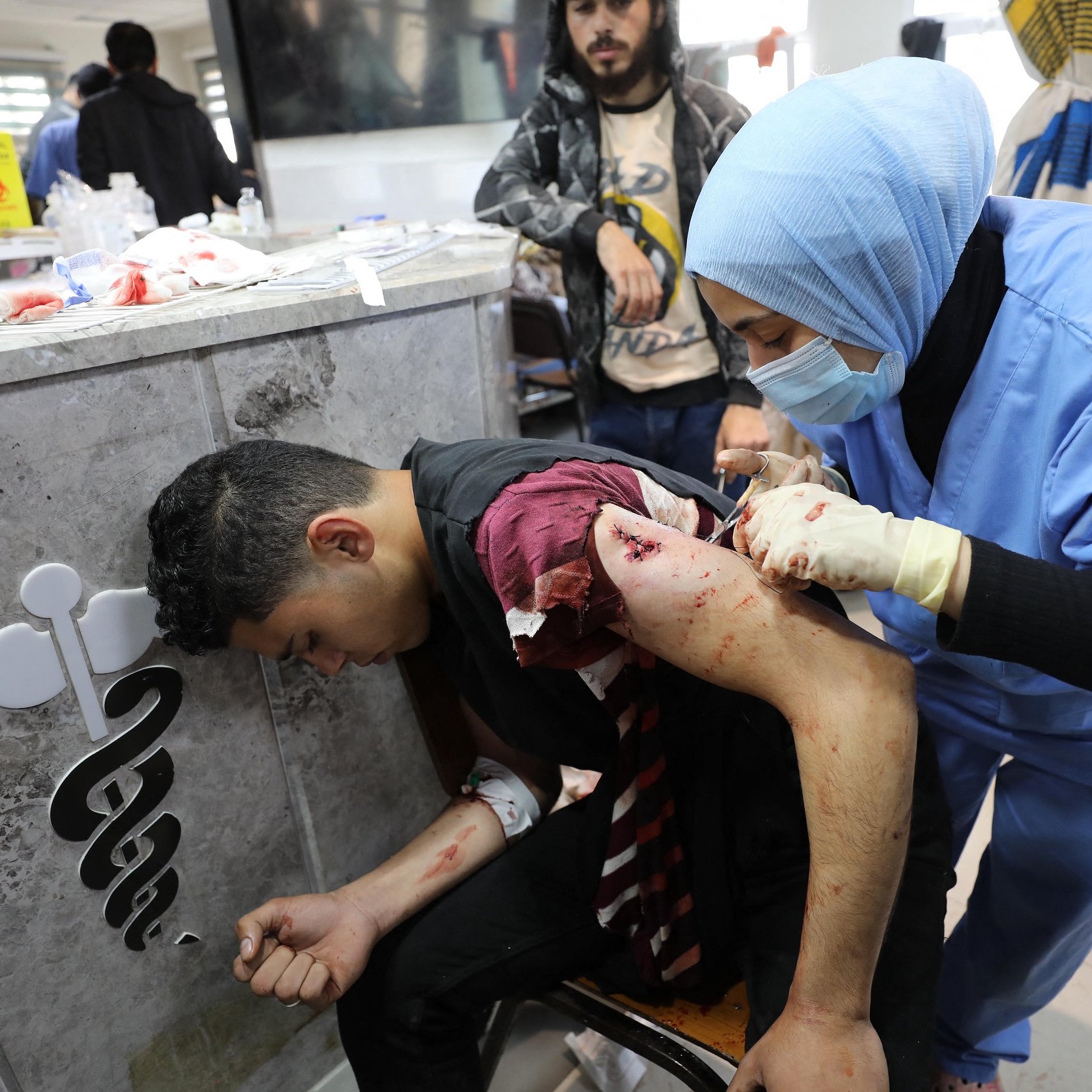 L'immagine mostra una scena in un ambiente sanitario. Un giovane uomo, visibilmente ferito, è seduto su una sedia mentre una professionista sanitaria, vestita con una divisa blu e con guanti, sta medicando una sua ferita sul braccio. Il suo volto mostra preoccupazione, e l'atmosfera appare tesa. Sullo sfondo si possono vedere altre persone e attrezzature mediche, suggerendo un contesto di emergenza.