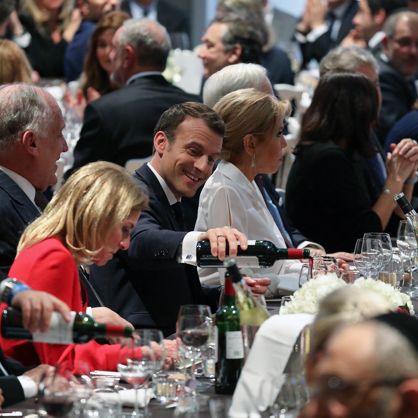 تصویر یک ضیافت یا مراسم رسمی را نشان می‌دهد که در آن افراد زیادی در کنار یکدیگر نشسته‌اند. در وسط تصویر، مردی با لباس رسمی و لبخند بر لب در حال سرو نوشیدنی به خانمی است که در سمت چپ او نشسته است. دیگر شرکت‌کنندگان نیز در اطراف میز حضور دارند و بعضی از آنها در حال گفتگو یا گوش دادن به صحبت‌های دیگران هستند. دکوراسیون میز با گل‌ها و شمع‌ها تزئین شده است و ظروف و لیوان‌های شفاف نیز روی میز قرار دارند. فضای تصویر به نظر دوستانه و گرم می‌آید.