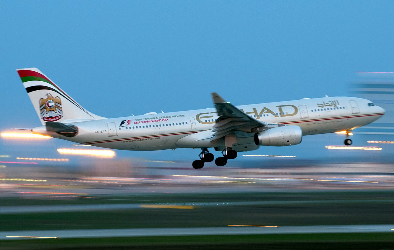 L'image montre un avion de la compagnie aérienne Etihad Airways en phase de décollage ou d'atterrissage. L'avion est partiellement flou, ce qui suggère une vitesse élevée. Le ciel apparaît dans une teinte bleue, tandis que des lumières brillantes en arrière-plan ajoutent une ambiance dynamique. L'appareil affiche la décoration et le logo caractéristiques de la compagnie.
