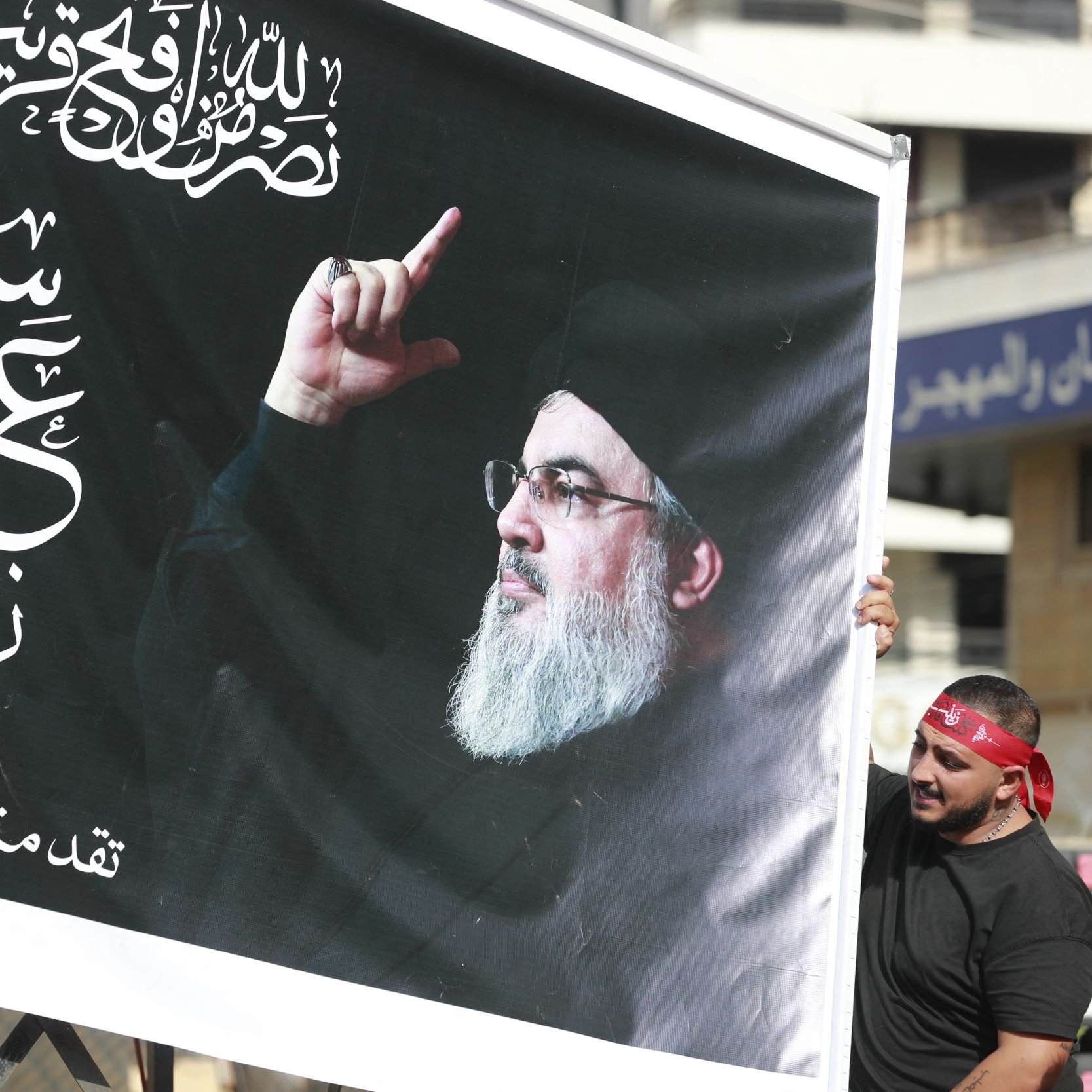 تصویر دو نفر را نشان می‌دهد که یک تابلو بزرگ را با تصویری از فردی در حال اشاره کردن به سمت بالا در دست دارند. پس‌زمینه تابلو سیاه است و بر روی آن متن‌هایی به زبان عربی دیده می‌شود. افراد حاضر در تصویر لباس‌های سیاه دارند و برخی از آن‌ها دستبندهای همرنگ بر روی سرشان دارند. این صحنه به نظر می‌رسد که در یک تجمع یا مراسم خاصی در حال برگزاری است.