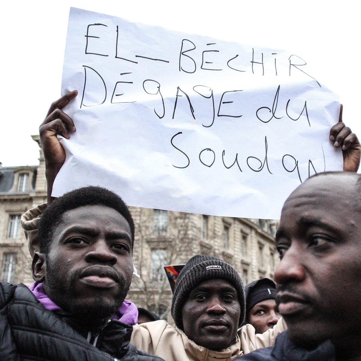 L'image montre une manifestation où des personnes tiennent des pancartes. L'une d'elles est clairement visible et affiche un message en français disant "El-Béchir dégage du Soudan". Les participants semblent passionnés et engagés dans une cause. On peut discerner des expressions sérieuses et déterminées sur leurs visages. En arrière-plan, des bâtiments peuvent être aperçus, suggérant un cadre urbain.