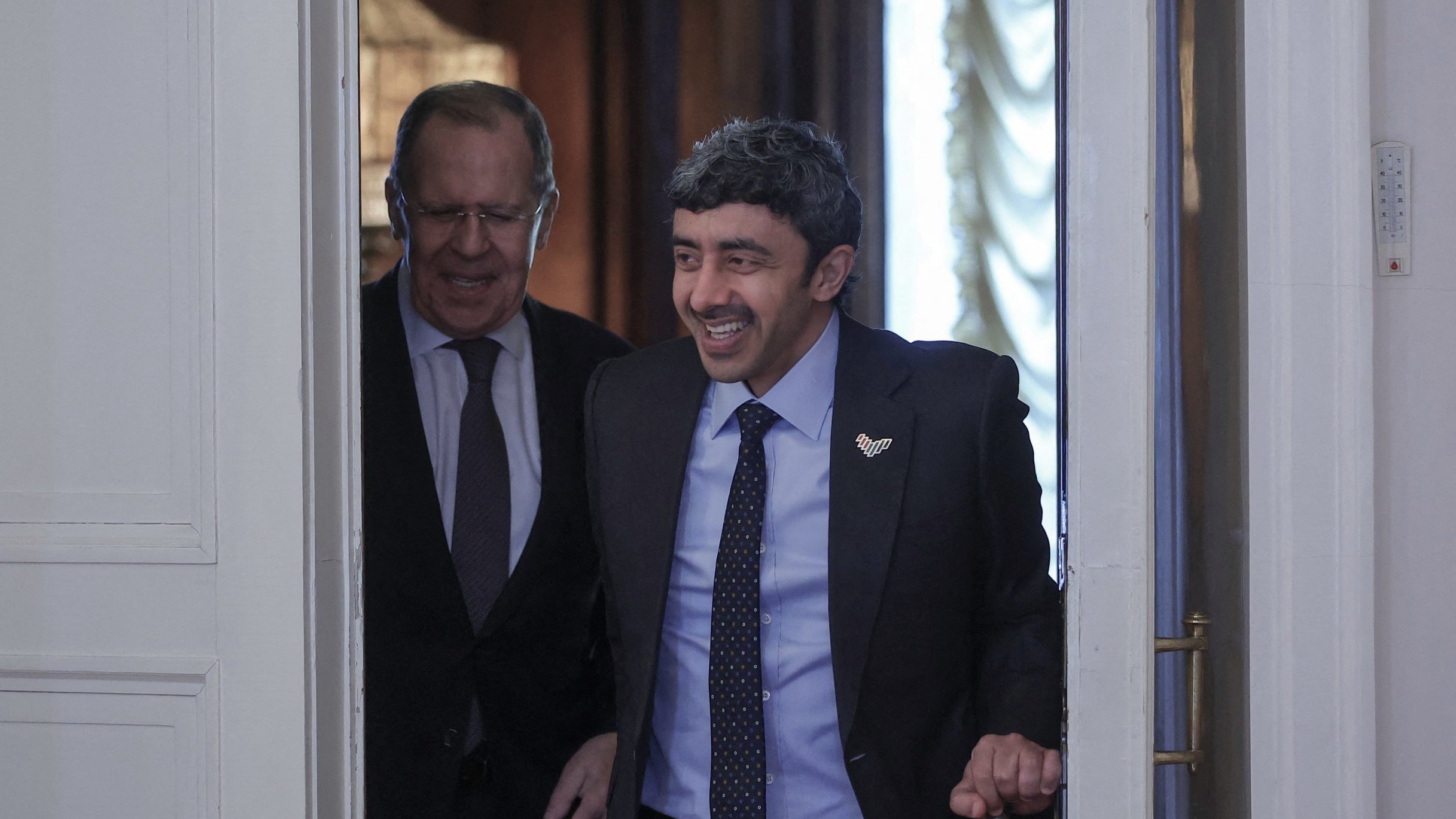 تظهر الصورة شخصين يتحدثان أثناء دخولهما من باب. الشخص في المقدمة يبدو مبتسمًا ويبدو وكأنه في حالة مزاجية جيدة، بينما الشخص الآخر خلفه يقدم تعبيرًا ودودًا. الخلفية تحتوي على تفاصيل معمارية تبرز الطابع الرسمي للمكان. كلا الشخصين يرتديان بدلات رسمية، مما يشير إلى حدث دبلوماسي أو اجتماع مهم.