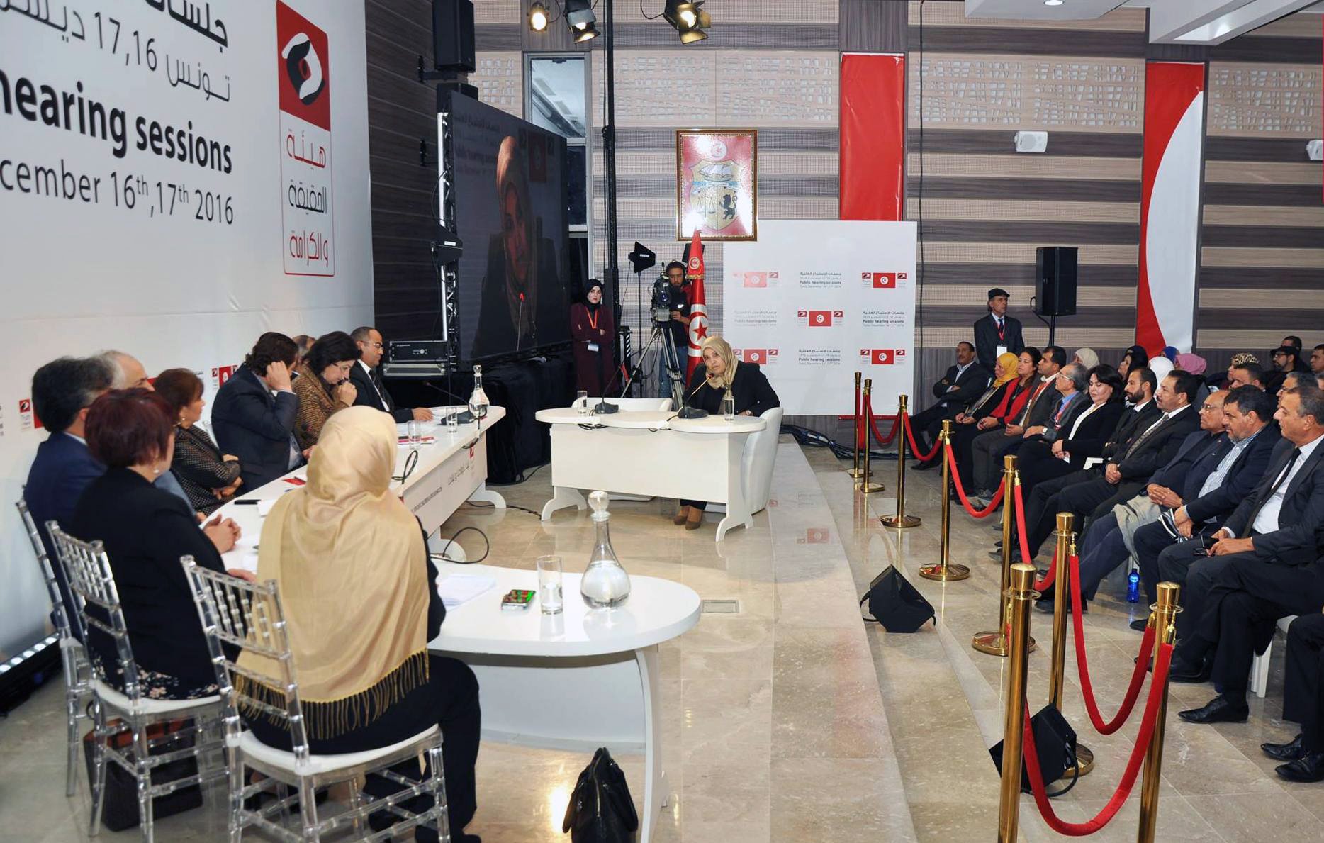الصورة تظهر جلسة استماع تتعلق بموضوع اجتماعي أو سياسي. في المكان، يجلس عدد من الأشخاص على الجانبين، حيث يبدو أن هناك مجموعة من المتحدثين يجلسون على منصة مرتفعة. يظهر أمامهم طاولة مستديرة حيث يتم تقديم المداخلات. تُظهر الخلفية شعارات وصورة لمتحدث ما، مما يشير إلى أهمية الحدث. الأجواء تبدو رسمية ومرتبة، مع وجود حواجز حمراء تفصل بين الحضور.