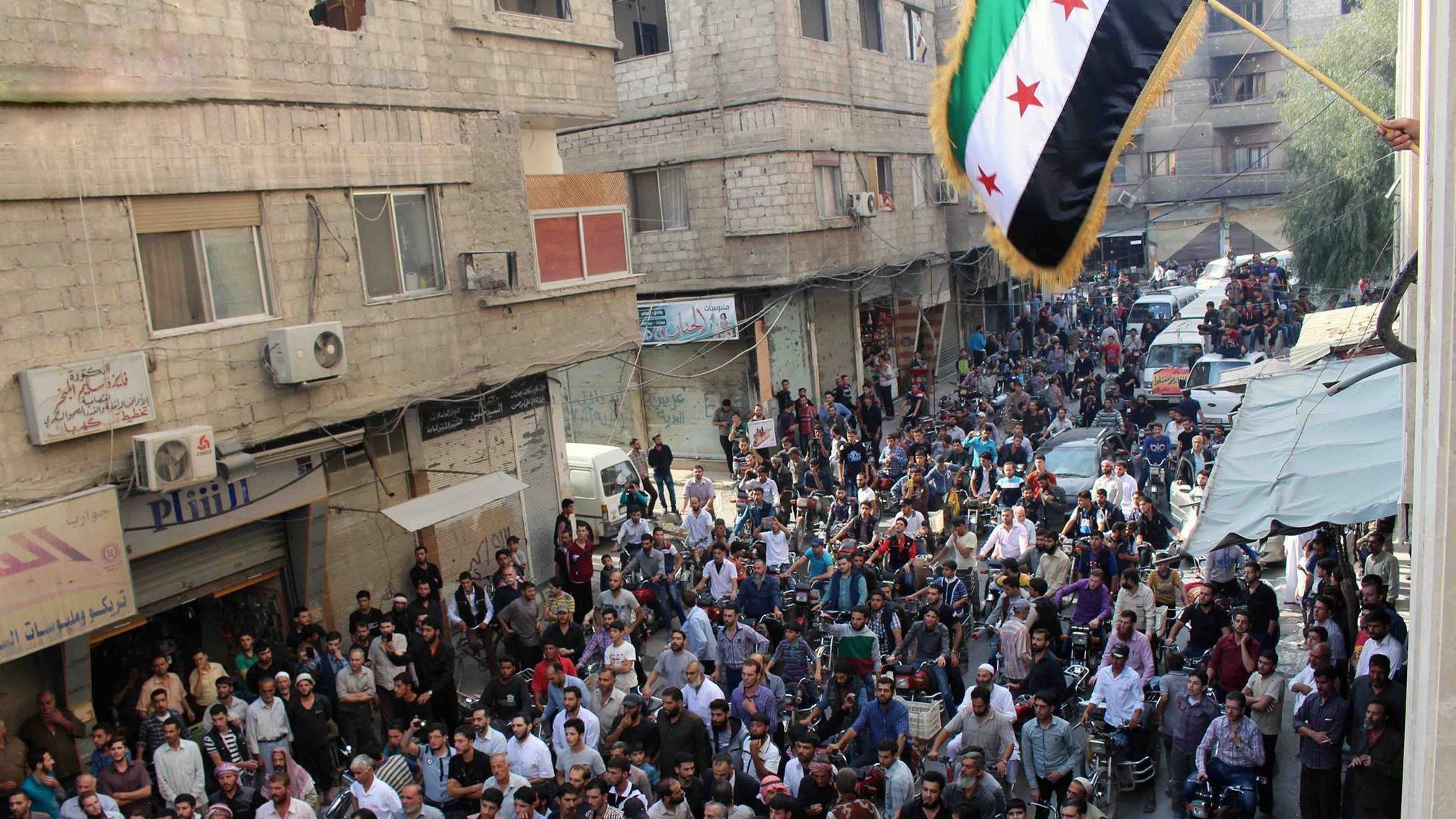 الصورة تظهر حشودًا كبيرة من الناس في شارع مزدحم. يتجمع المحتجون في منطقة حضرية، حيث يمكن رؤية المباني والأسطح والشرفات. علم يرفرف في الأعلى، وهو العلم السوري المعتمد من قبل بعض فصائل المعارضة. يبدو أن هناك جو من الحماس والتجمع، مع وجود بعض السيارات والمركبات في الخلفية.