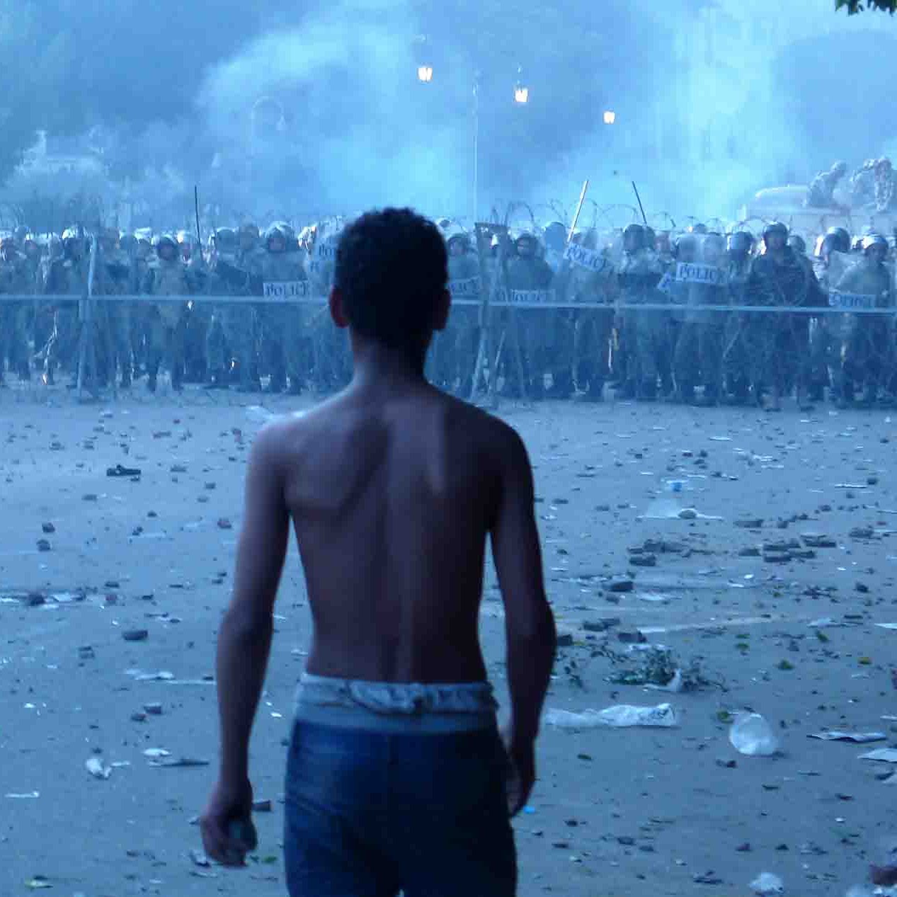 تصویر نشان‌دهنده یک صحنه تنش‌زا است. در جلو، یک پسر جوان با تنه برهنه به سمت گروهی از نیروهای انتظامی که در فاصله‌ای دورتر ایستاده‌اند، نگاه می‌کند. این نیروها در لباس‌های نظامی و با سپرهایی در دست، آماده مواجهه هستند. زمین در اطراف پر از زباله و بقایای ناآرامی‌هاست، و بخاری یا دودی در هوا وجود دارد که جو پرتنش‌تری را ایجاد می‌کند. فضای کلی تصویر احساس اضطراب و کشمکش را منتقل می‌کند.