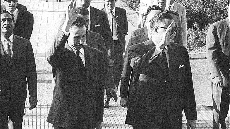 The image depicts a group of men walking together outdoors in a formal setting. Two men in the foreground are waving, suggesting they are acknowledging an audience or photographers. They are dressed in suits, indicating a formal occasion. Behind them, several other men in suits and uniforms follow, along with some individuals in lighter clothing, possibly indicating a recent gathering or event. The background features greenery and is well-organized, contributing to a dignified atmosphere.