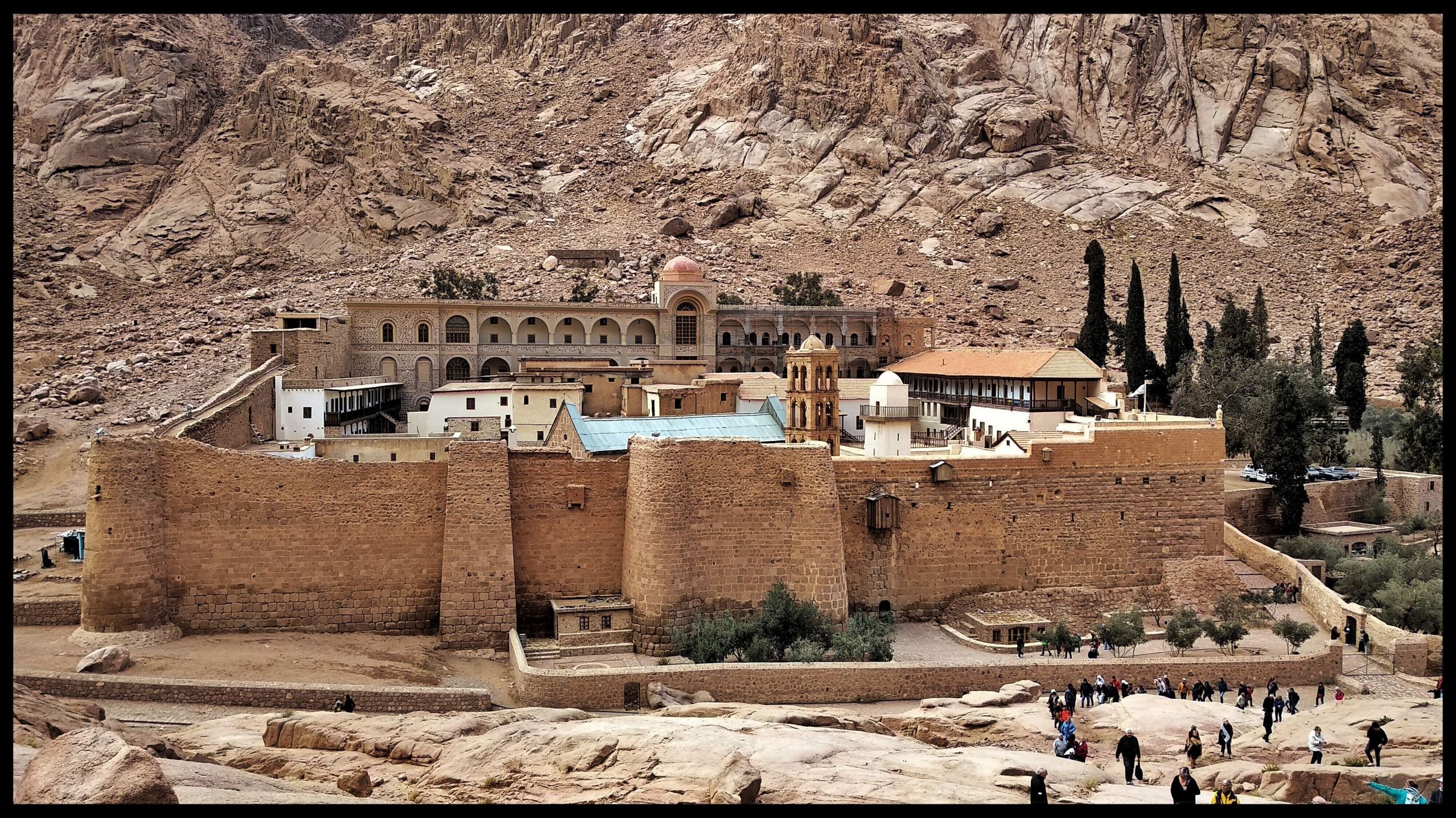 L'image montre un monastère situé dans une région montagneuse aride. Le bâtiment, de style architectural ancien, est construit en pierre et possède des murs épais. On peut voir une grande cour intérieure ainsi que des arbres qui ajoutent une touche de verdure au paysage. À l'arrière-plan, on distingue des formations rocheuses spectaculaires, soulignant l'isolement et la beauté naturelle des lieux. Des visiteurs se promènent autour du monastère, ce qui témoigne de son attrait touristique.