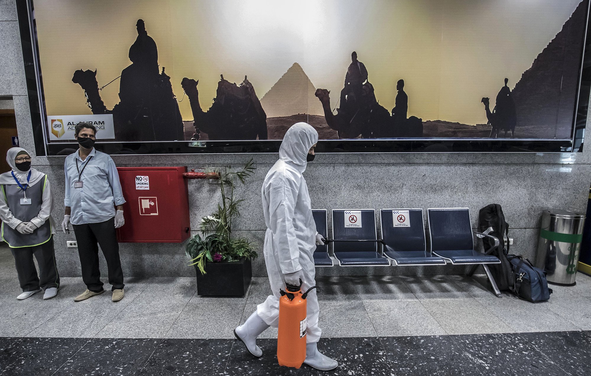 L'image montre un hall d'aéroport ou une station, avec un fond représentant des silhouettes de chameaux et de pyramides, visiblement au coucher du soleil. Au premier plan, une personne en costume de protection blanc marche, tenant une valise orange. Deux personnes en uniforme, probablement du personnel de sécurité ou de santé, se tiennent sur le côté, portant des masques. L'environnement est moderne et propre, avec des sièges en métal et des éléments de décoration comme des plantes.