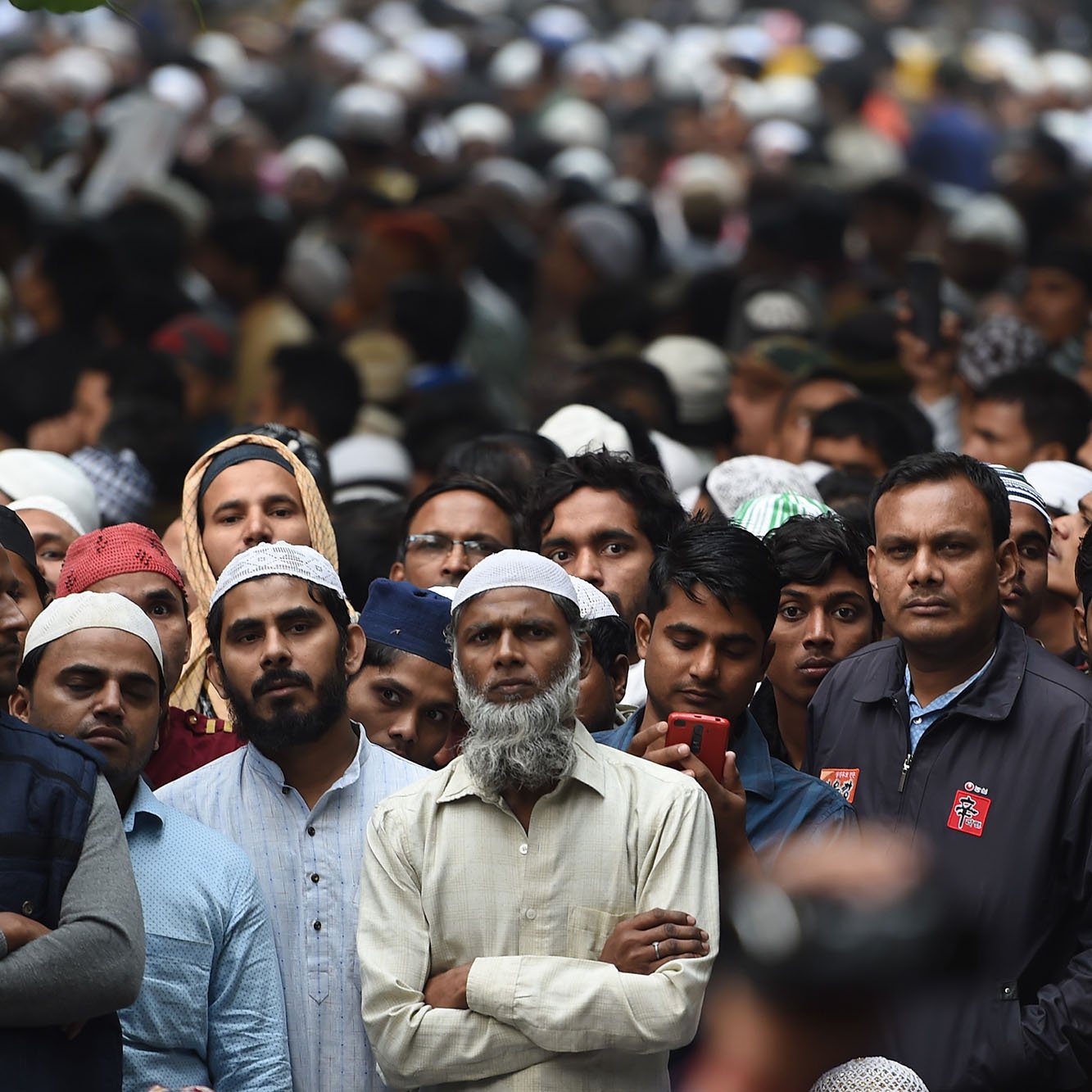 The image shows a large crowd of people, primarily men, gathered closely together. Many of the individuals are wearing traditional Islamic attire, including caps (taqiyah or kufi). The expressions on their faces suggest a mix of focus and anticipation. The background is filled with more attendees, creating a sense of a significant event or gathering. The atmosphere appears to be one of unity and collective participation.