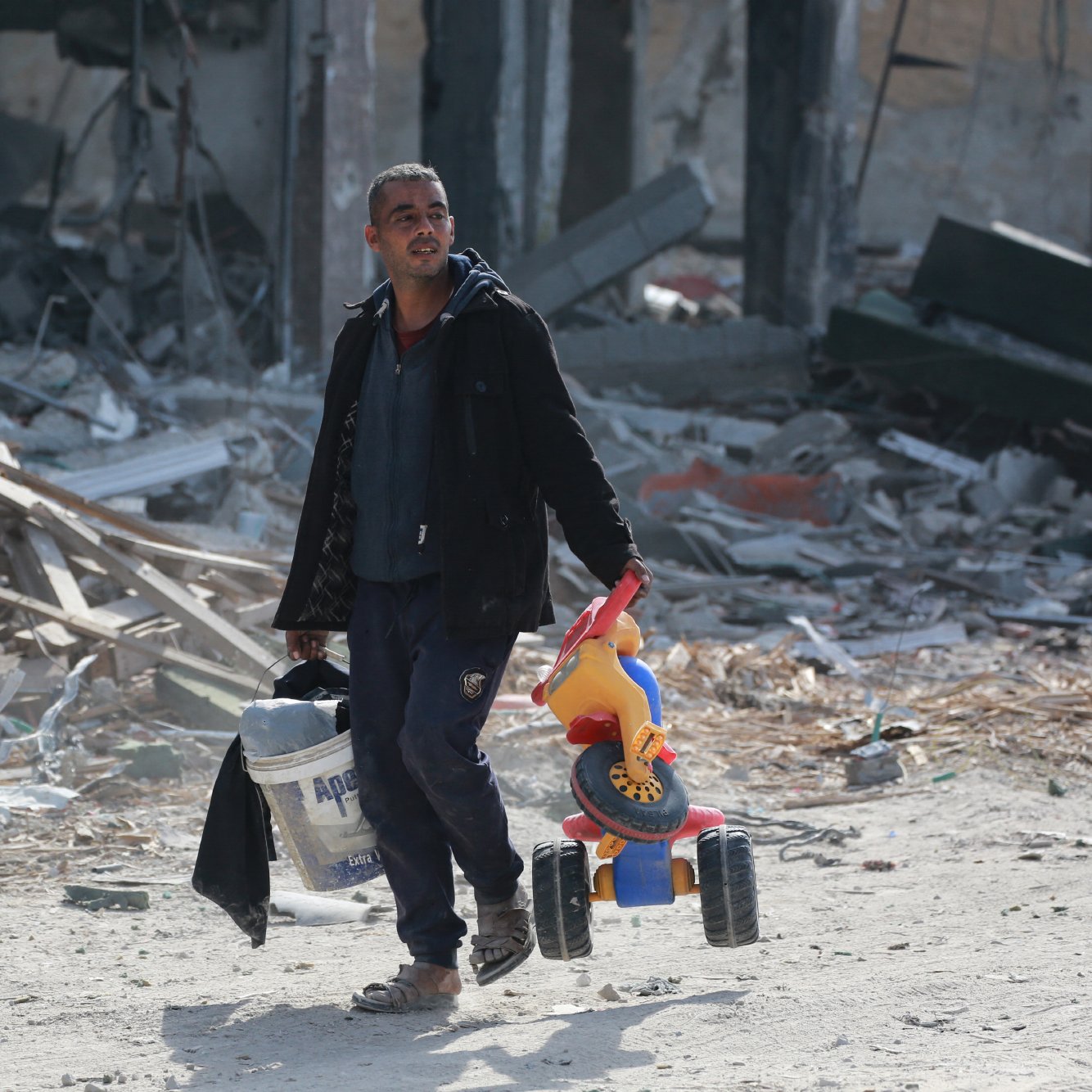 تصویر مردی را نشان می‌دهد که در فضایی ویران شده و پر از آوار حرکت می‌کند. او با ظاهری خسته، یک سطل در دست دارد و در دست دیگرش یک اسباب‌بازی رنگی که شبیه به دوچرخه است، حمل می‌کند. پس‌زمینه تصویر شامل بقایای ساختمان‌ها و مواد ساختمانی پراکنده است که نمایانگر وضعیت ناراحت‌کننده‌ای است. 