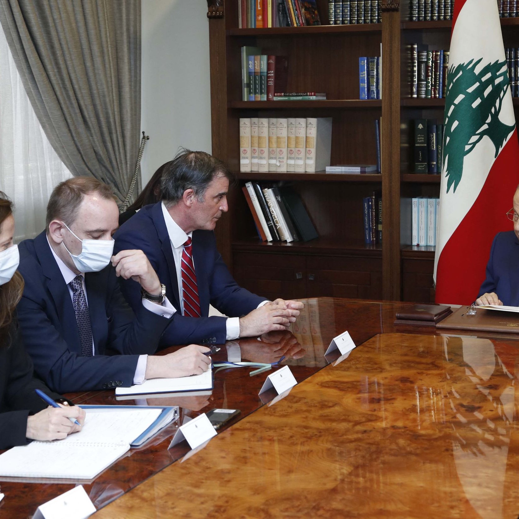 Nell'immagine, si può vedere un incontro ufficiale in una sala ben arredata. Ci sono quattro persone sedute attorno a un grande tavolo in legno. A destra, un uomo anziano, presumibilmente un leader o un ufficiale governativo, è rivolto verso gli altri partecipanti. Gli altri tre, seduti a sinistra, indossano mascherine e stanno prendendo appunti su quaderni. Dietro di loro, si intravede una libreria piena di libri e una bandiera del Libano. L'atmosfera sembra seria e professionale, tipica di una discussione di Stato.