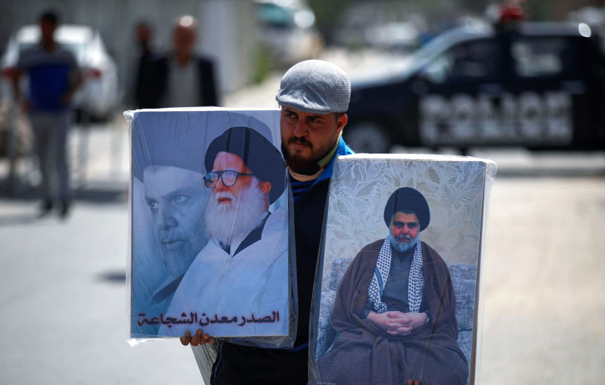 L'immagine mostra un uomo che trasporta due grandi ritratti. A sinistra si vede un'immagine di un leader religioso, mentre a destra c'è un altro ritrato di un diverso leader. L'uomo indossa un berretto e sembra camminare in un contesto urbano, con un veicolo della polizia visibile sullo sfondo. L'atmosfera indica una manifestazione o un evento che riguarda temi religiosi o politici.