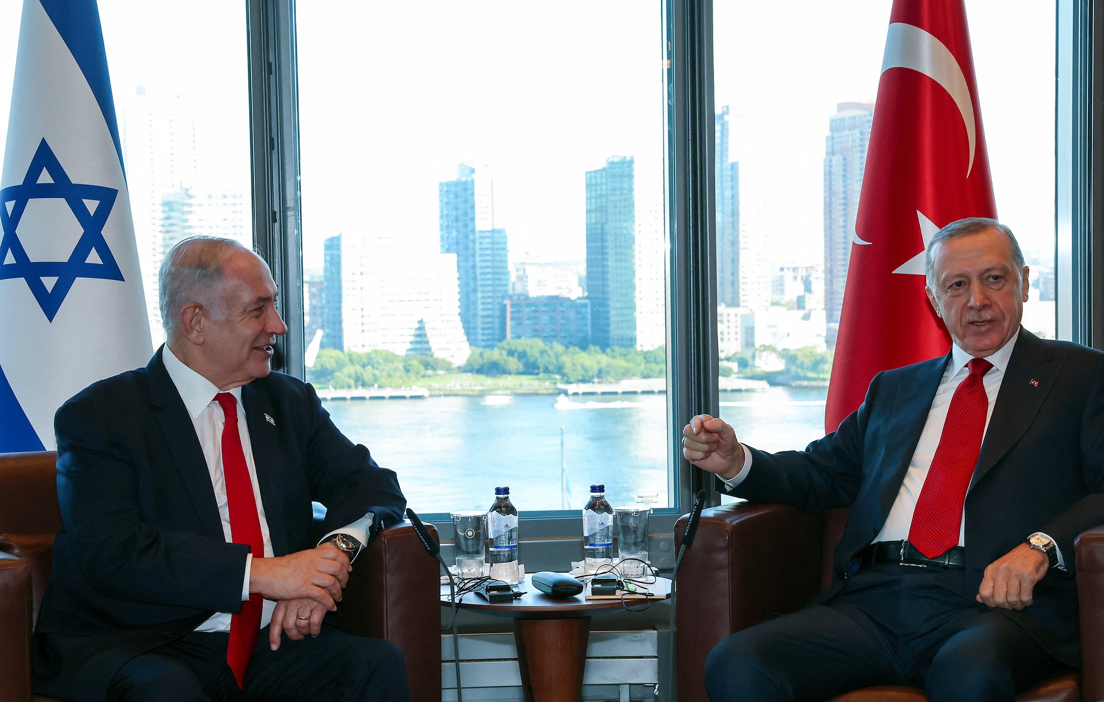 این تصویر دو فرد را در حال گفت‌وگو نشان می‌دهد. در سمت چپ، پرچم اسرائیل و در سمت راست، پرچم ترکیه قرار دارد. این افراد در یک اتاق با نمایی از شهر در پس‌زمینه نشسته‌اند که ساختمان‌های بلند و دریاچه‌ای را نشان می‌دهد. هر دو نفر به نظر می‌رسد که در حال تبادل نظر دوستانه هستند. 