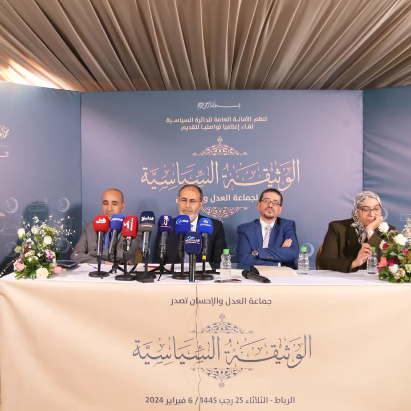 الصورة تظهر مؤتمرًا صحفيًا أو اجتماعًا رسميًا. يوجد على المنصة أربعة أشخاص يجلسون معًا، حيث يتحدث أحدهم ويبدو أنه يتحدث في ميكروفون. هناك لافتة خلفهم تحمل عنوان الاجتماع، ومجموعة من الكاميرات والشاشات تسجل أو تنقل الحدث. الطاولة مزينة بالزهور، والإضاءة هادئة. الخلفية تبدو أنيقة وتعكس الجدية الرسمية للاجتماع.