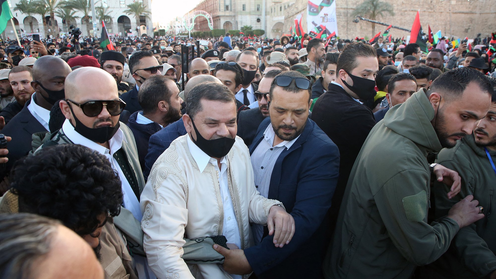 الصورة تُظهر حشودًا كبيرة من الناس في مكان عام، بعضهم يرتدي الكمامات، ويبدو أن هناك شخصيات بارزة في الوسط. يظهر الأشخاص في الصورة ممالأة وتفاعل مع بعضهم، مما يوحي بأن هناك حدثًا مهمًا يجري. يمكن أن يكون الحدث مظاهرة أو تجمعًا سياسيًا، حيث تحمل الأعلام وتظهر بعض الوجوه المألوفة. الجو بشكل عام يبدو متوترًا ولكن مليئًا بالحماس.