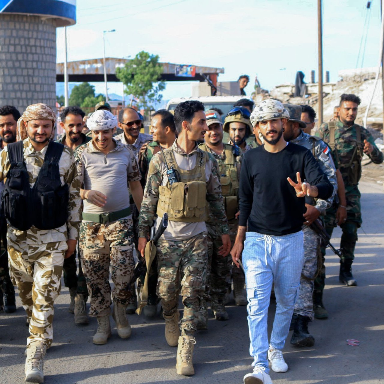 La imagen muestra a un grupo de hombres, aparentemente soldados, caminando juntos por una calle. Algunos llevan uniformes militares y portan armas, mientras que otros visten ropa más casual. Hay un ambiente de camaradería, y uno de los hombres en el centro hace un gesto de paz con la mano. El fondo muestra un paisaje urbano y estructuras que parecen ser parte de una ciudad en reconstrucción o conflicto. La escena refleja un momento de unidad y posiblemente celebración.