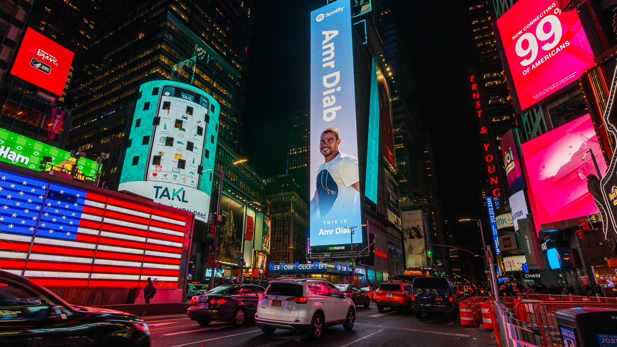 L'image montre une scène animée de Times Square à New York, illuminée par de nombreux panneaux publicitaires lumineux. Au centre de l'image, on peut voir une grande affiche mettant en avant une personne, Amr Diab, avec un sourire. Les bâtiments environnants sont recouverts de publicités colorées, dont l'une représente un drapeau américain. On aperçoit également des voitures circulant dans la rue, ajoutant à l'ambiance dynamique de ce célèbre endroit. Les lumières vives et les écrans créent une atmosphère vibrante et énergique.