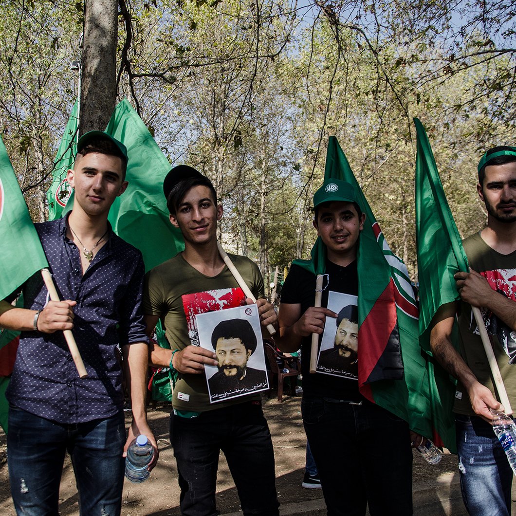 L'image montre un groupe de quatre jeunes hommes rassemblés lors d'un événement, probablement une manifestation ou une célébration. Ils tiennent des drapeaux verts, qui semblent avoir un symbole spécifique. Chacun d'eux porte des affiches représentant des figures, et ils affichent des expressions de fierté ou de détermination. L'arrière-plan est verdoyant, évoquant un cadre festif en plein air.