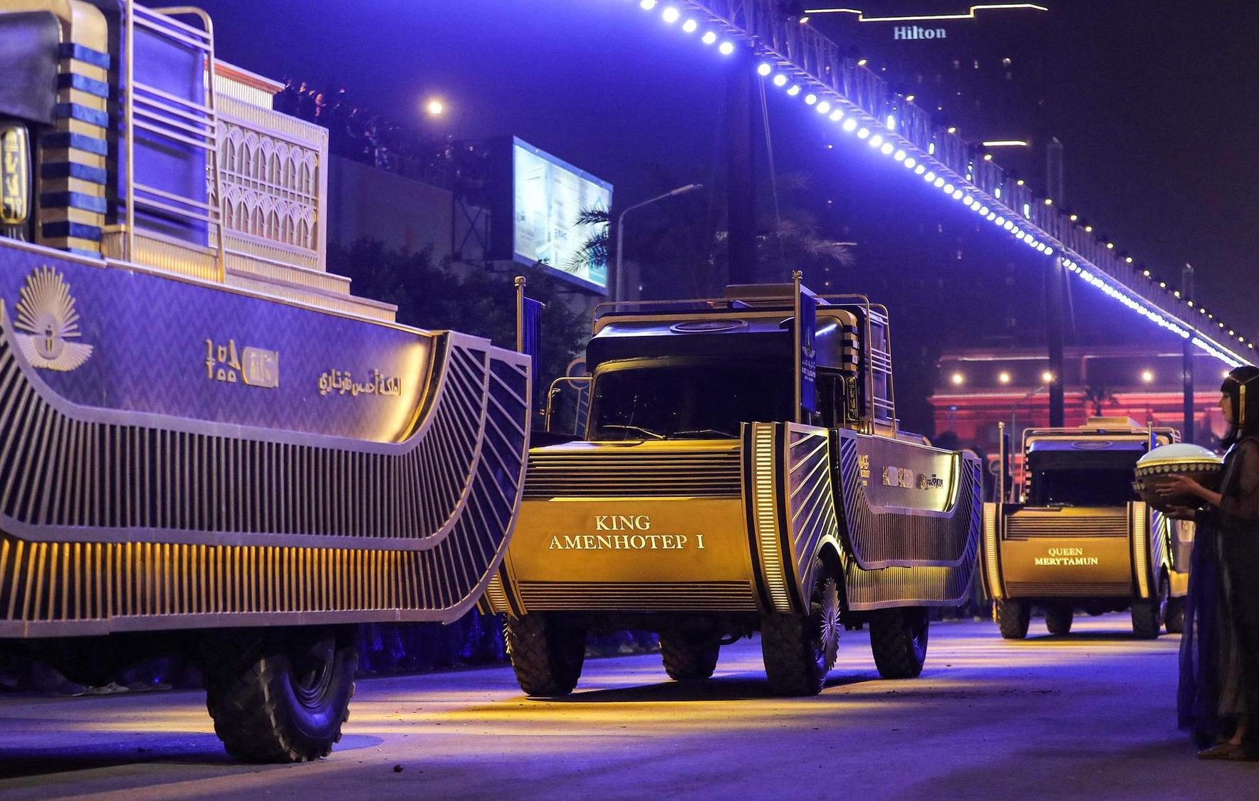 La imagen muestra una escena nocturna de un desfile en la que varios camiones decorados están alineados en una calle iluminada. Los camiones tienen un diseño llamativo y parecen estar relacionados con temas históricos, específicamente sobre reyes egipcios, como "KING AMENHOTEP I". En el primer plano, hay una persona vestida con ropa tradicional que sostiene un objeto, posiblemente una ofrenda o un instrumento musical. El ambiente es festivo y majestuoso, con luces brillantes que realzan el ambiente del evento. En el fondo, se pueden observar edificios y carteles, lo que sugiere que el desfile está teniendo lugar en una ciudad moderna.