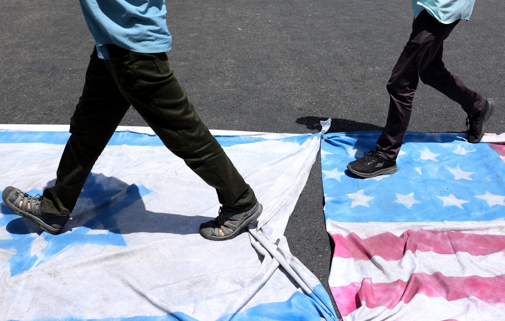 دو نفر در حال قدم زدن روی پرچم‌های دو کشور هستند. زمین زیر آن‌ها خاکی و تاریک است.