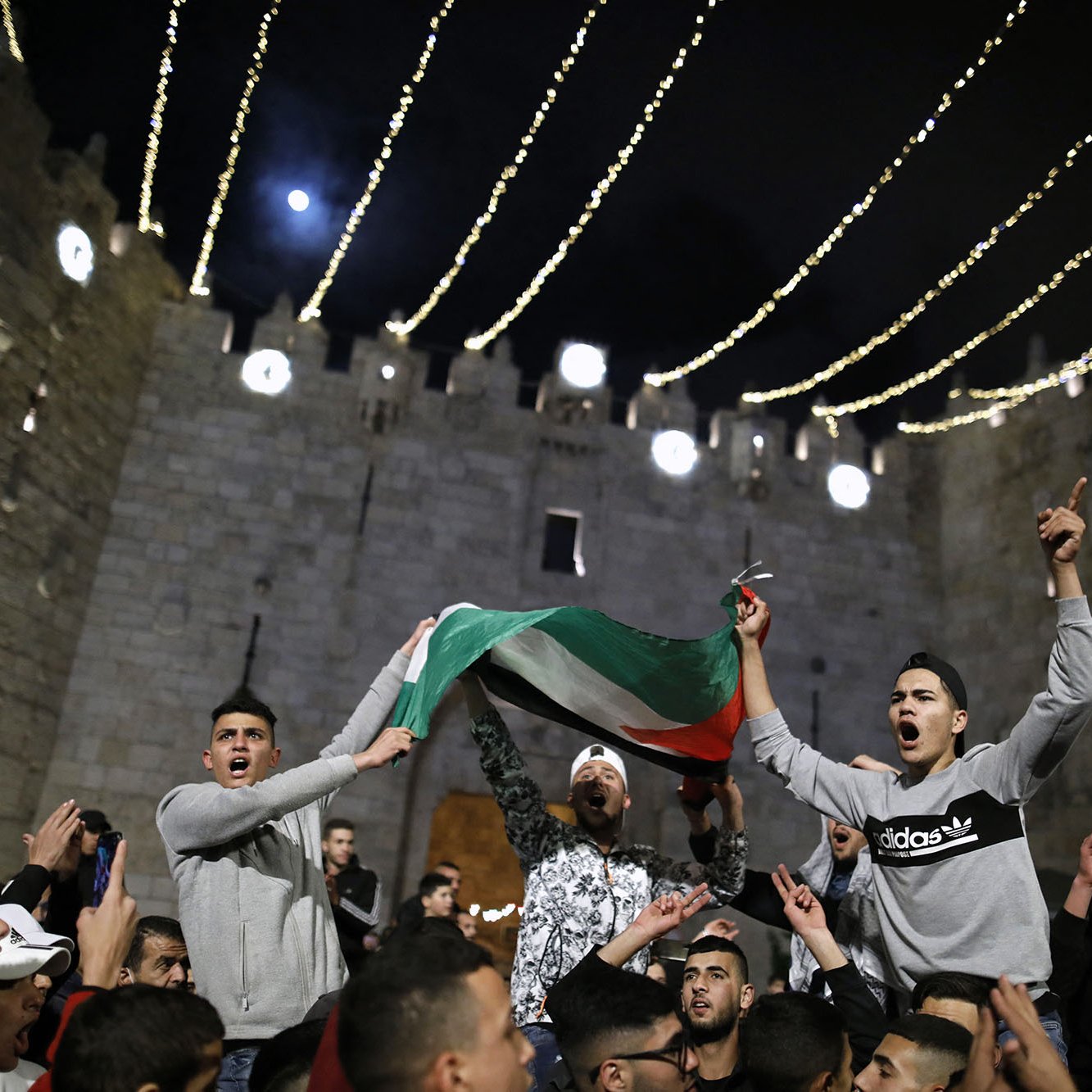 L'immagine mostra un gruppo di persone riunite in un contesto festivo, probabilmente in un evento pubblico. I partecipanti stanno sventolando una bandiera, che sembra essere quella palestinese, e sono visibilmente entusiasti. Sullo sfondo, si può notare una struttura medievale, che suggerisce un'atmosfera di celebrazione. Le luci appese creano un'atmosfera suggestiva e notturna, mentre i volti delle persone esprimono gioia e partecipazione.