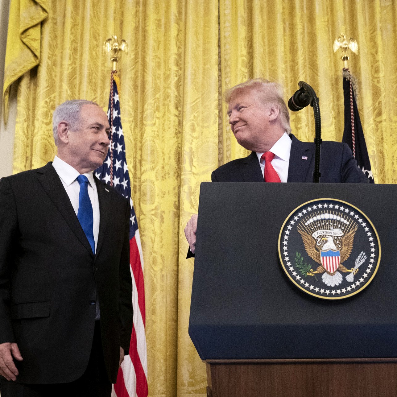 L'immagine mostra due uomini in un ambiente ufficiale, probabilmente in una cerimonia o conferenza stampa. Sono di fronte a un podio con il sigillo presidenziale degli Stati Uniti. Sullo sfondo, ci sono delle tende dorate e le bandiere statunitensi. I due uomini sembrano sorridere e interagire in modo cordiale.