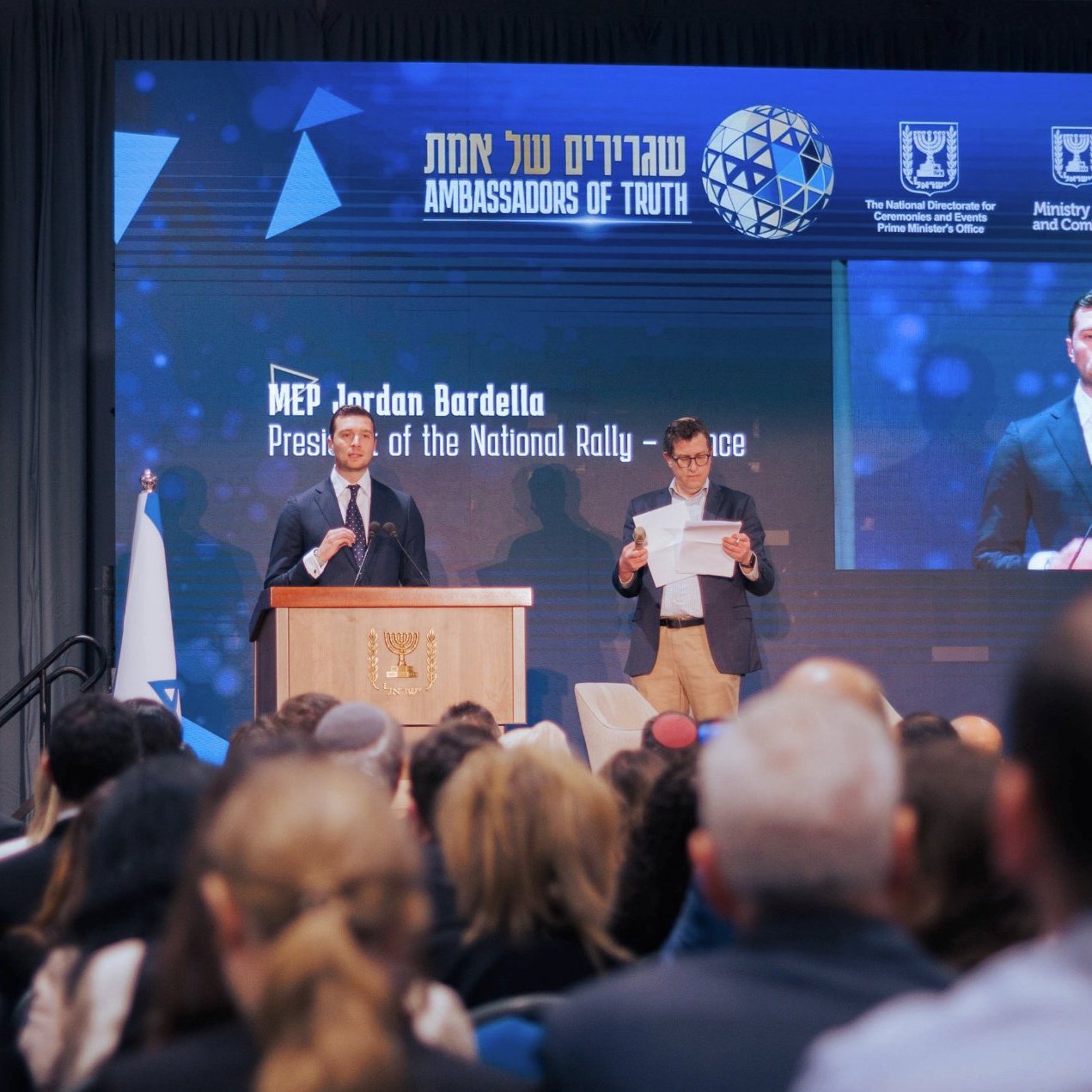 L'image montre une conférence avec un orateur debout derrière un pupitre, lisant un discours. Le public clairement visible est assis devant lui, et un grand écran derrière affiche des informations sur l'événement. L'atmosphère est formelle et engageante.