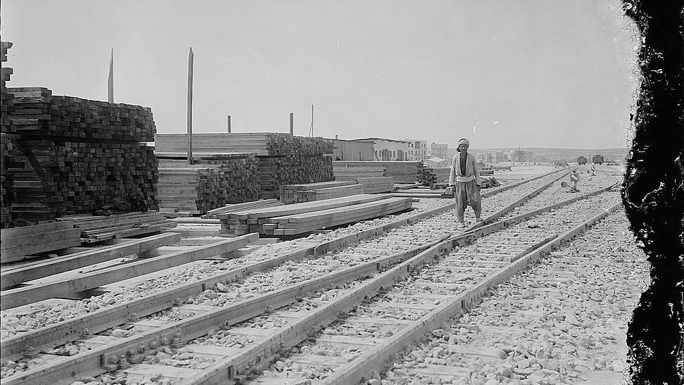 L'image montre une scène industrielle avec des voies de chemin de fer se dirigeant vers l'horizon. Sur le côté gauche, il y a des tas de planches de bois empilées, ce qui suggère une activité de construction ou de stockage. Un homme, probablement un travailleur, marche le long des rails. En arrière-plan, on aperçoit des bâtiments, donnant l'impression d'un site en pleine activité. Le ciel est dégagé, et la lumière est vive, typique d'une journée ensoleillée. L'atmosphère générale évoque une époque passée, avec une ambiance de travail acharné.