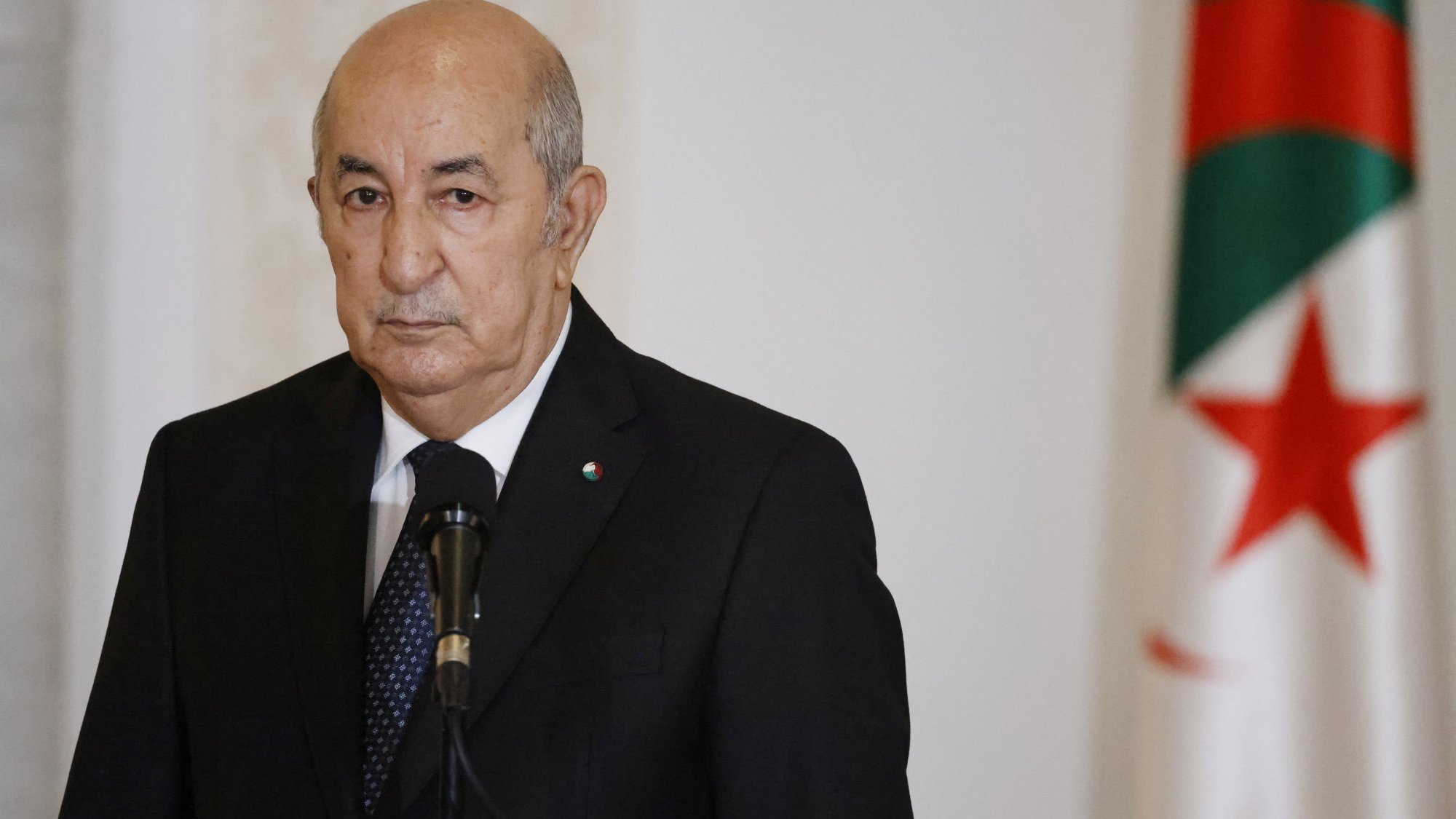 The image depicts a middle-aged man dressed in a dark suit, standing at a podium with a microphone. He has a serious expression on his face. In the background, there is an Algerian flag featuring a green and white field with a red star and crescent. The setting appears to be an official environment, likely a government or press conference area.