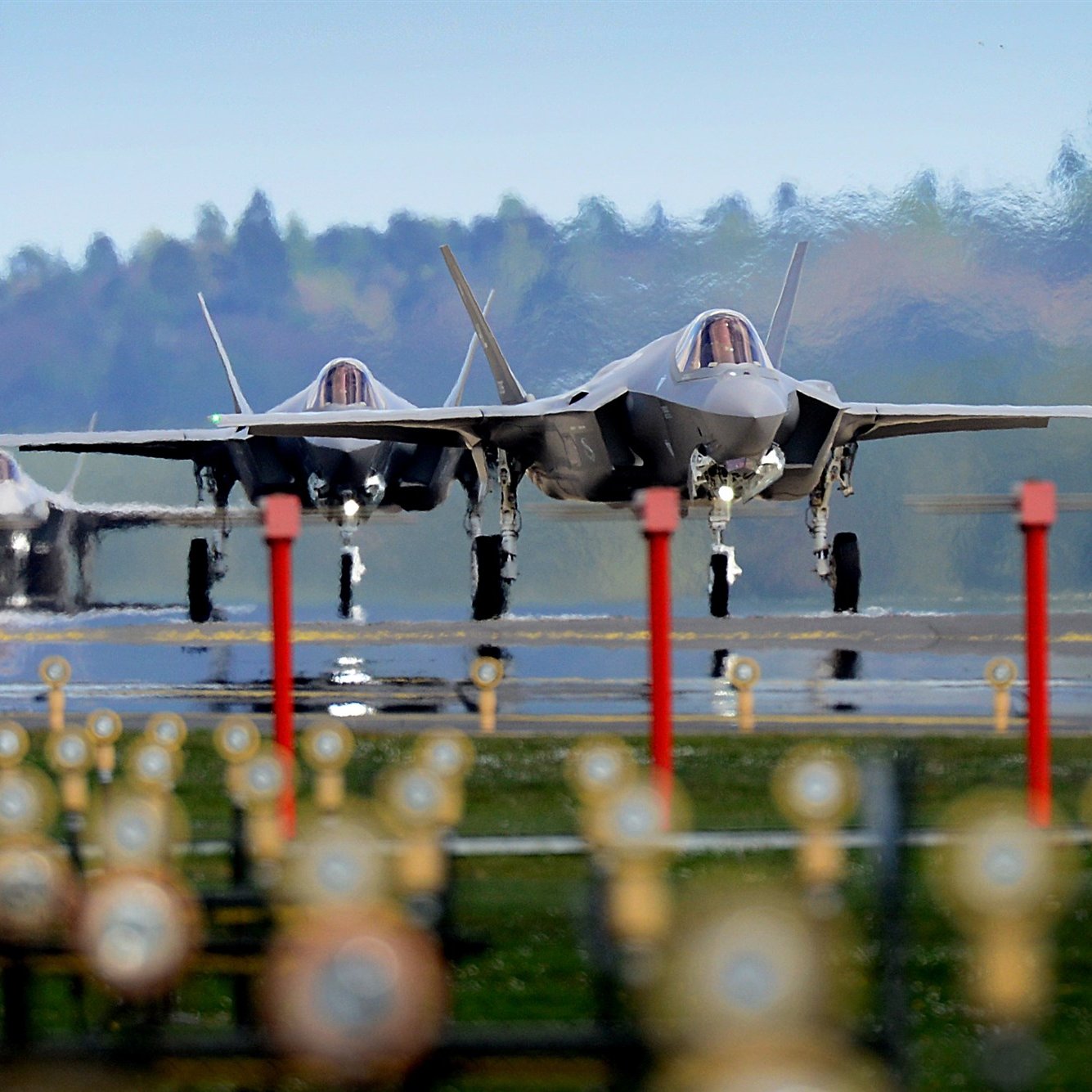 The image features three F-35 fighter jets on an airstrip, showcasing their sleek and modern designs. The jets are positioned closely together, and the background features a blurred green landscape, indicating a rural setting. The foreground includes several lights lining the runway, which adds a sense of depth to the scene. The overall atmosphere appears dynamic and focused on military aviation.