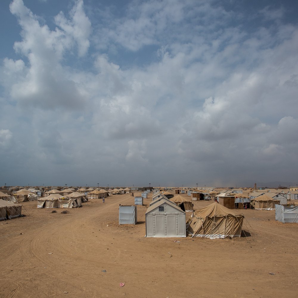 L'image montre un paysage désertique avec des tentes disposées dans une vaste étendue de sable. Le ciel est nuageux, avec des nuages gris et blancs qui donnent une ambiance tranquille mais assez chargé. Les tentes sont de couleurs variées, majoritairement beige et grise, et semblent être organisées de manière assez dense, suggérant qu'il s'agit d'un camp, probablement pour des personnes déplacées ou réfugiées. L'environnement général est aride et peu végétalisé, accentuant l'aspect isolé du lieu.