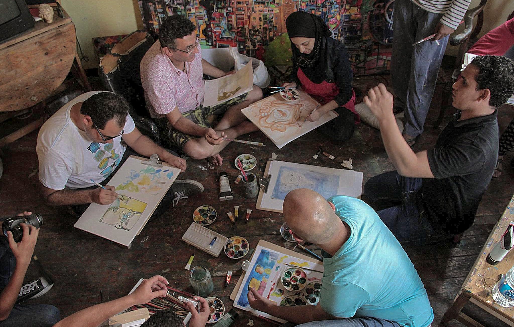 تظهر في الصورة مجموعة من الأشخاص يجلسون على الأرض في غرفة مليئة بالألوان واللوحات. يعملون على رسم لوحات فنية باستخدام فرش وألوان مختلفة، حيث يبدو أن بعضهم يركز على تفاصيل رسوماته بينما يتحدث الآخرون. هناك أيضًا مجموعة من الألوان والفرش والأدوات الفنية موضوعة حولهم. أجواء الصورة تعكس روح التعاون والإبداع في الفن.