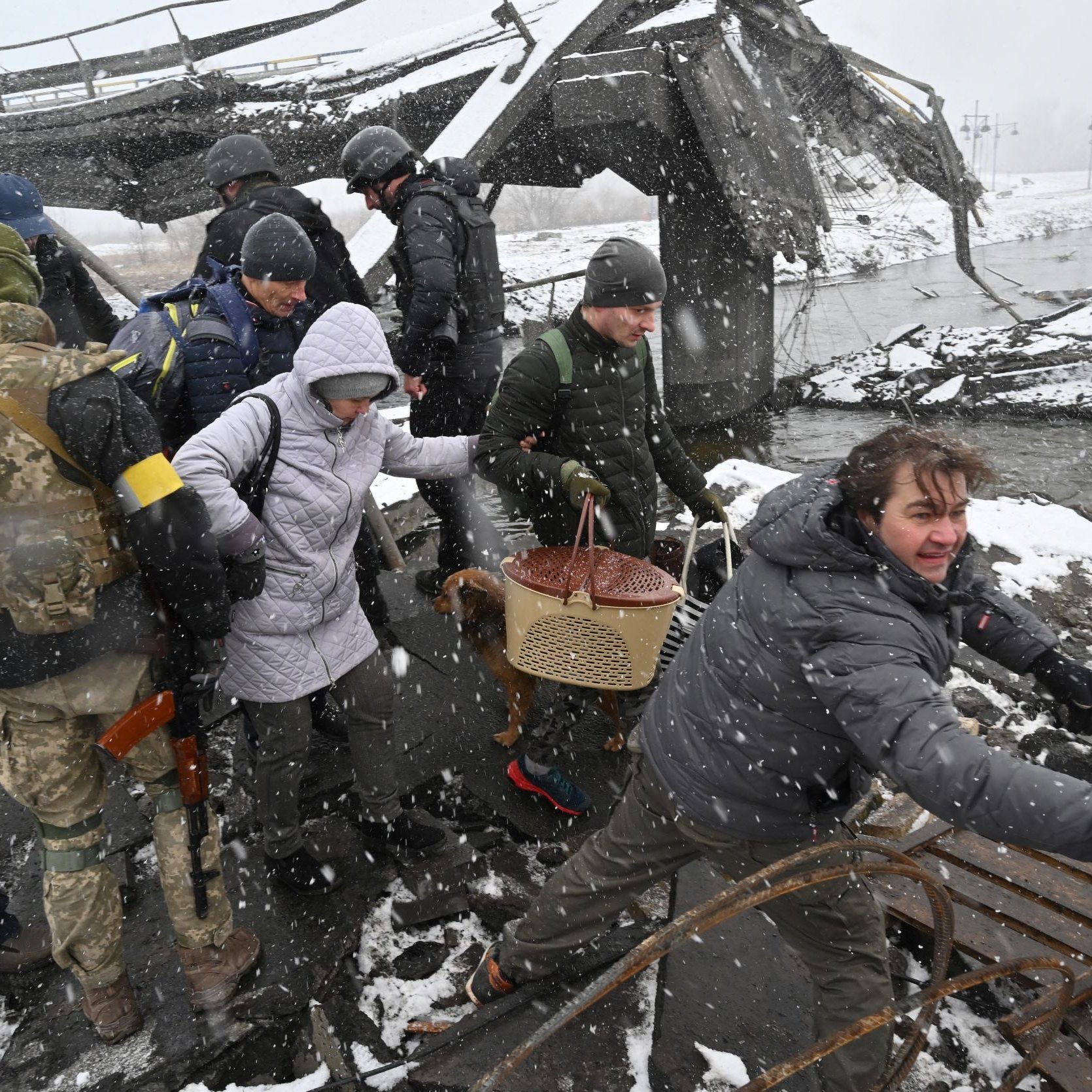 L'immagine mostra un gruppo di persone in un'area distrutta, probabilmente a causa di un conflitto o di un disastro. Alcune persone indossano abbigliamento militare, mentre altre sembrano civili. È in corso un salvataggio o un'evacuazione, con qualcuno che sta aiutando un'altra persona a attraversare il terreno difficile. La scena è caratterizzata da neve e condizioni atmosferiche difficili, suggerendo un ambiente critico e di emergenza. I volti mostrano preoccupazione e determinazione.