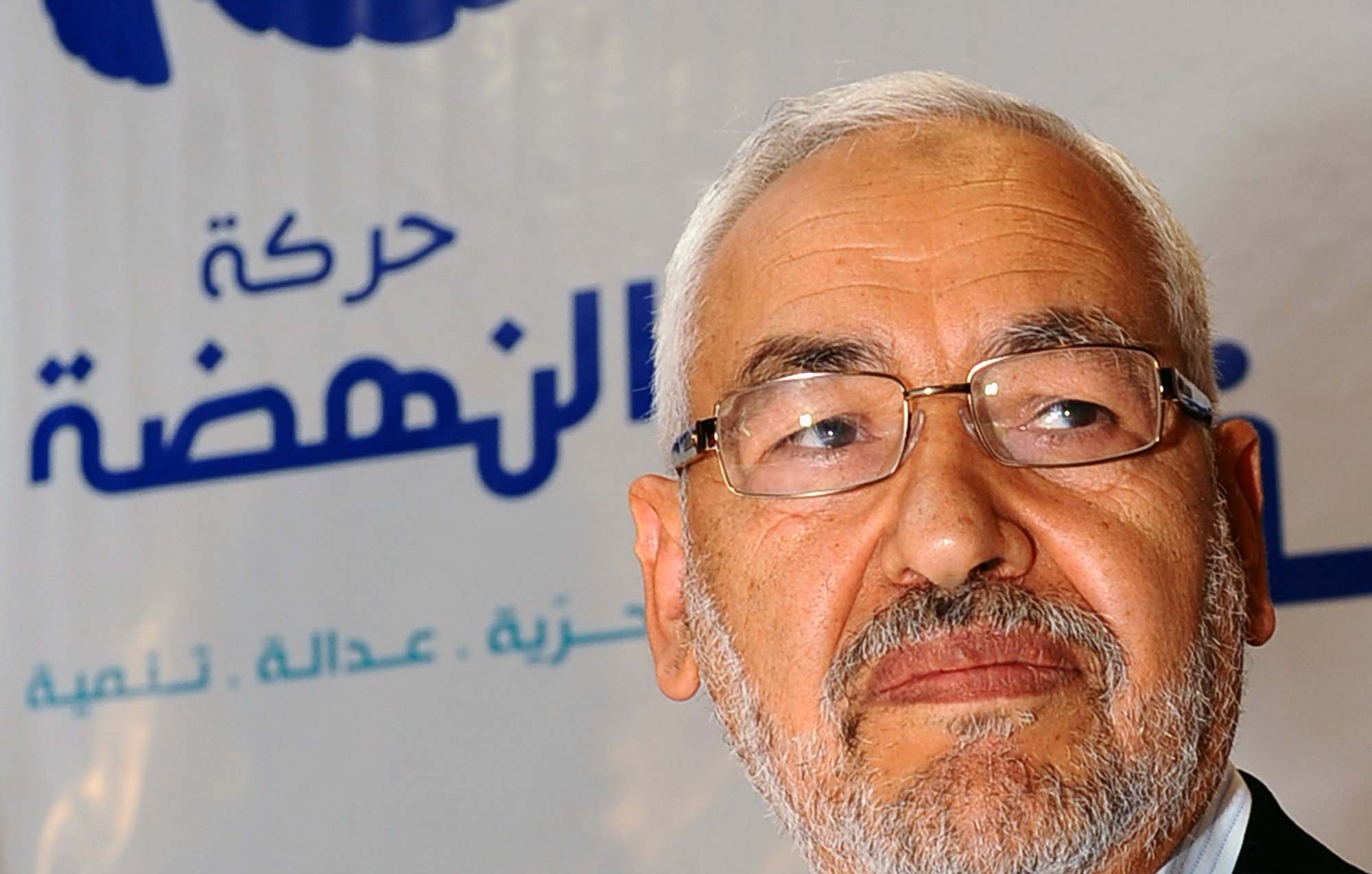 الصورة تظهر شخصًا يرتدي نظارات ويملك لحية وشعرًا أبيض، يبدو وكأنه يتحدث أو يعبر عن رأي. خلفه شعار يحمل اسم "حركة النهضة" والذي يشير إلى حزب سياسي في تونس. تظهر الألوان في الخلفية بشكل بسيط، مما يركز الانتباه على الشخص في المقدمة.