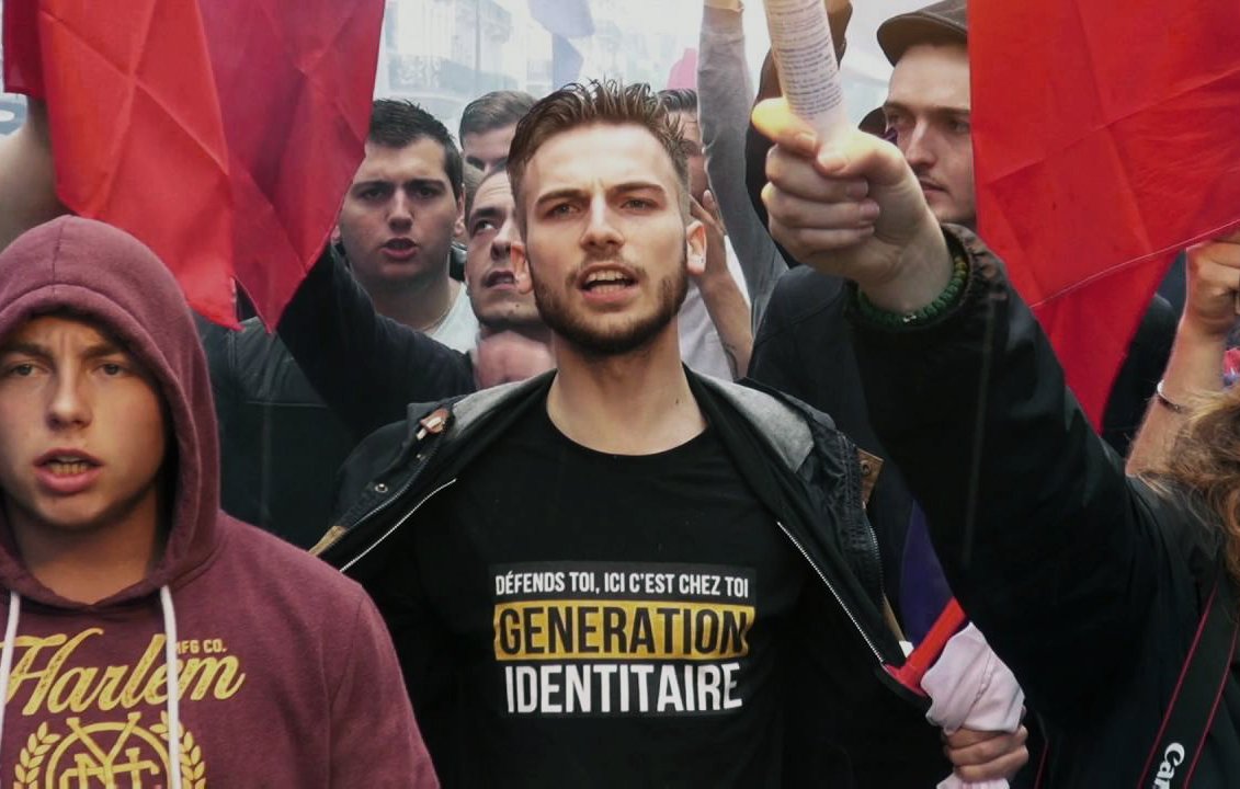 The image depicts a protest or demonstration scene. In the foreground, a young man stands prominently, wearing a black shirt with the text "GÉNÉRATION IDENTITAIRE" printed in yellow. He appears to be leading the group, holding a piece of paper in one hand and raising it in the air. Surrounding him are several other individuals, some of whom are holding red flags. The atmosphere seems charged and passionate, with many faces visible, indicative of a collective movement. The overall context suggests a rally focused on identity or nationalist themes.