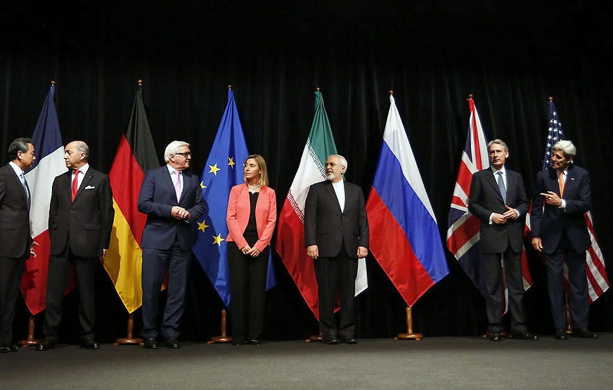 تظهر الصورة مجموعة من القادة أو الممثلين السياسيين واقفين أمام مجموعة من الأعلام لدول مختلفة، تشمل الصين، ألمانيا، الاتحاد الأوروبي، إيران، روسيا، وبريطانيا. يبدو أن الصورة قد تم التقاطها خلال حدث رسمي، حيث يتحدث بعضهم بينما ينظر الآخرون بشكل جاد. الأعلام مرتبة بشكل منظم، مما يعكس سياق سياسي دولي أو تفاوضي.