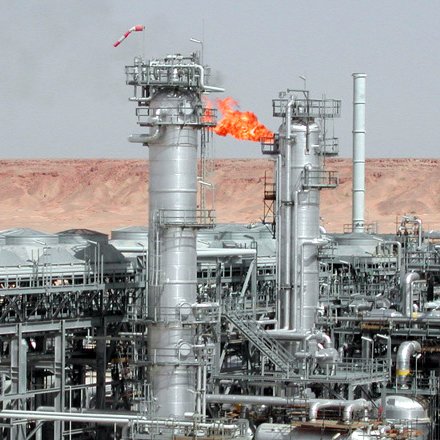 L'image montre une installation industrielle, probablement liée à l'industrie pétrolière ou gazière. On peut voir plusieurs tours métalliques, dont certaines émettent des flammes, indiquant une combustion, peut-être de gaz résiduel. L'arrière-plan présente un paysage désertique, caractérisé par des collines ou des montagnes sèches, typique des zones où se trouvent des installations énergétiques. Les structures sont en métal et semblent sophistiquées, témoignant d'une technologie avancée utilisée dans cette industrie.