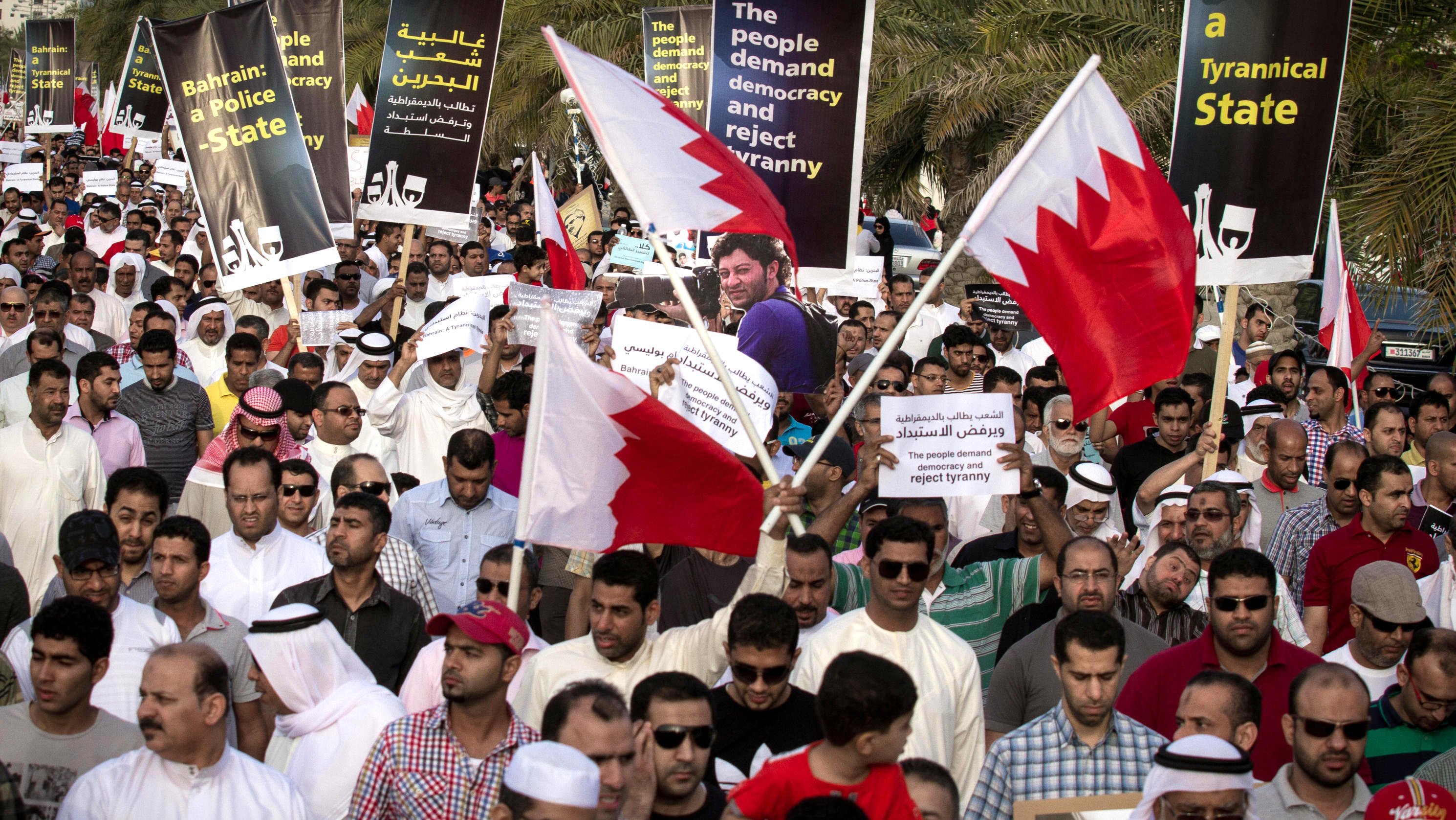 L'image montre une grande foule de manifestants rassemblés dans une rue, tenant des pancartes et des drapeaux de Bahreïn. Les manifestants semblent exprimer des revendications politiques, demandant la démocratie et critiquant le régime en place. On peut voir des gens de différentes origines et genres, tous unis dans leur volonté de changement, avec des expressions déterminées sur leurs visages. L'environnement témoigne d'un climat de mobilisation et de protestation collective.