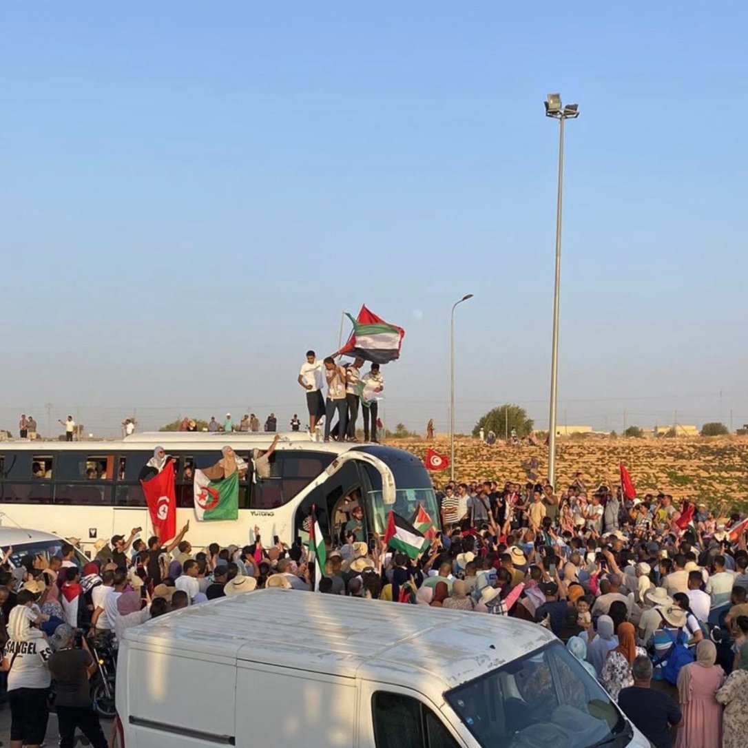 A large crowd gathers, waving flags and celebrating, with vehicles parked around.
