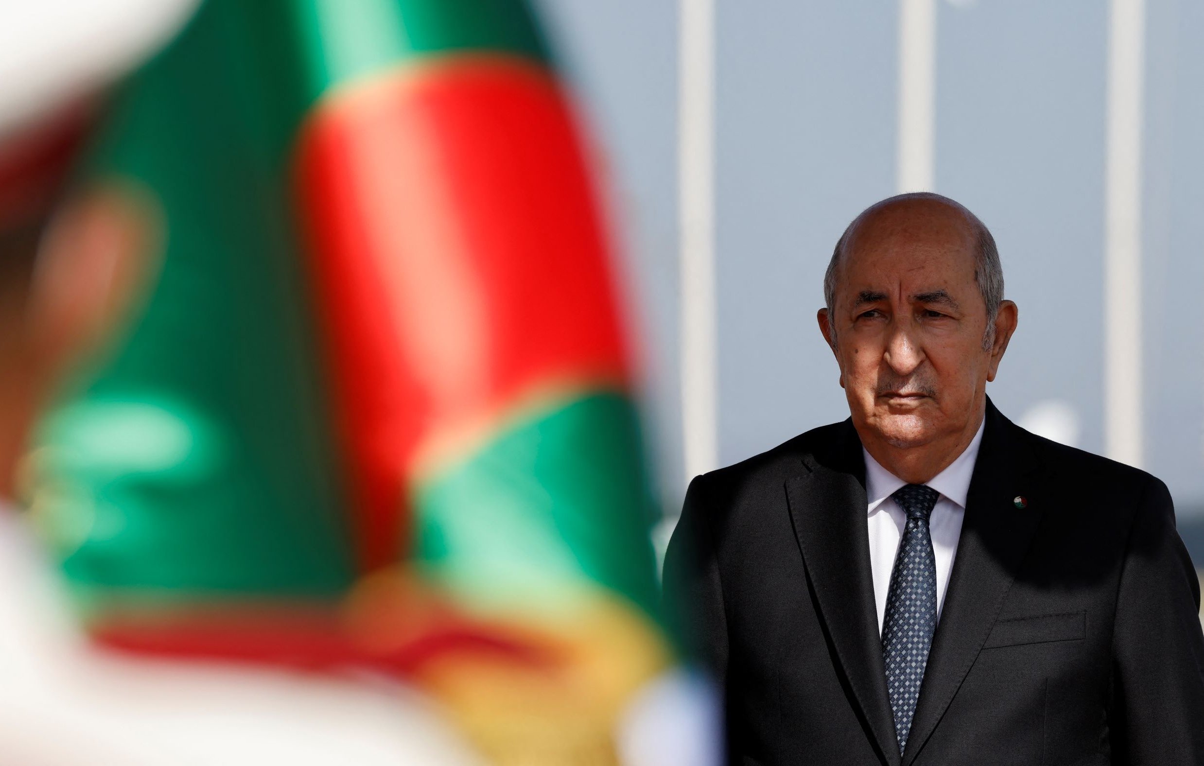 L'image montre un homme en costume sombre, se tenant droit avec une expression sérieuse, en arrière-plan d'un drapeau aux couleurs vives. Le flou devant l'homme suggère qu'il se trouve près d'une cérémonie officielle ou d'un événement important. Le drapeau vert et rouge crée un contraste avec son costume noir.