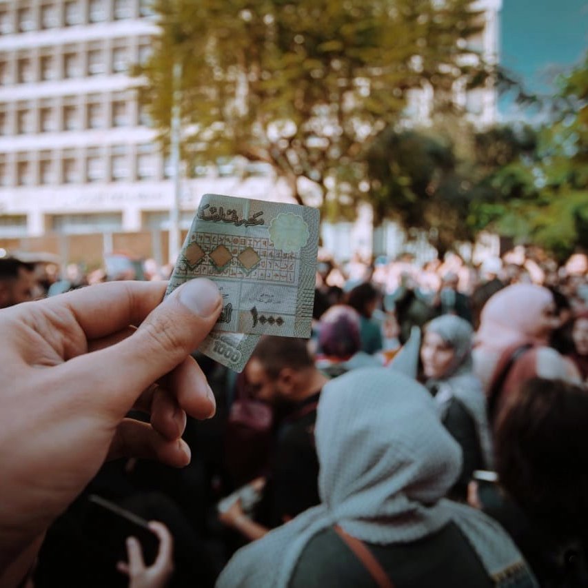 در این تصویر، یک دست در حال نگه‌داشتن یک کارت شناسایی یا مدرک مشابهی است. در پس‌زمینه، جمعیتی از افراد دیده می‌شود که در یک مکان عمومی جمع شده‌اند. برخی از افراد چادر یا پوشش‌های اسلامی دارند. نور روز و فضای خارجی نشان‌دهنده یک رویداد یا تجمع عمومی است. احساس عمومی شلوغی و فعالیت اجتماعی در این تصویر مشهود است.